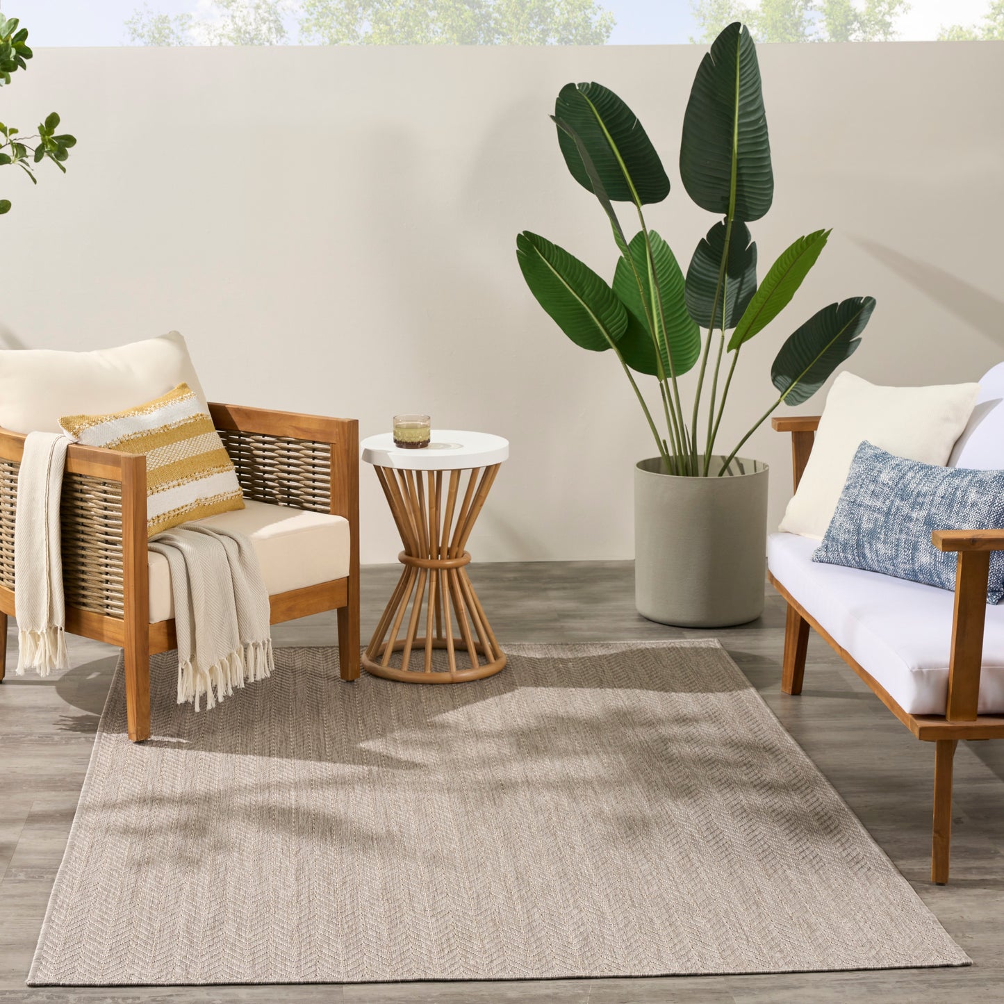Outdoor patio with wooden-framed armchairs, beige cushions, striped pillow, round white table, tall potted tropical plant.