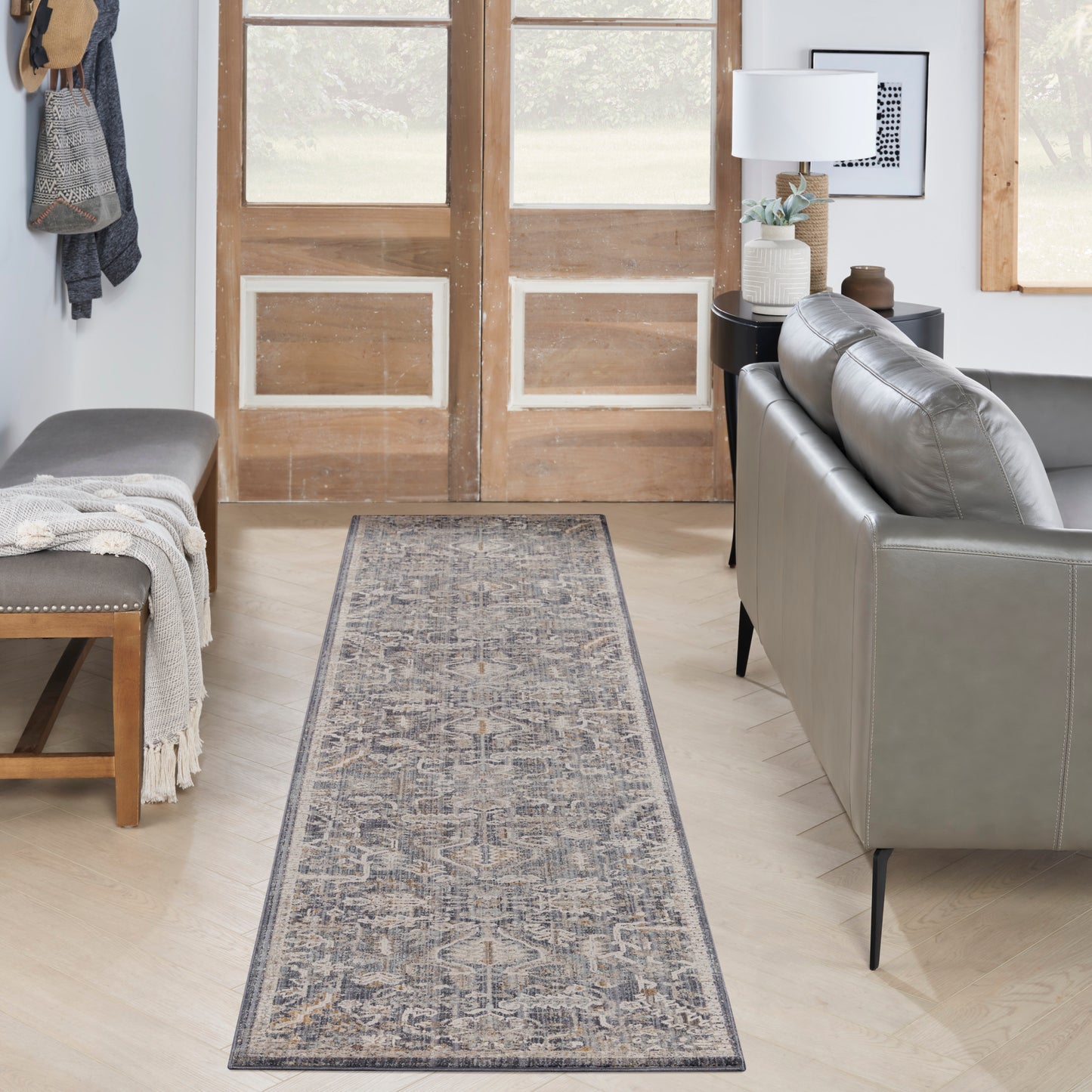 Blue-gray patterned runner rug on light wood floor in a cozy entryway with gray leather sofa, bench, & rustic wooden doors.