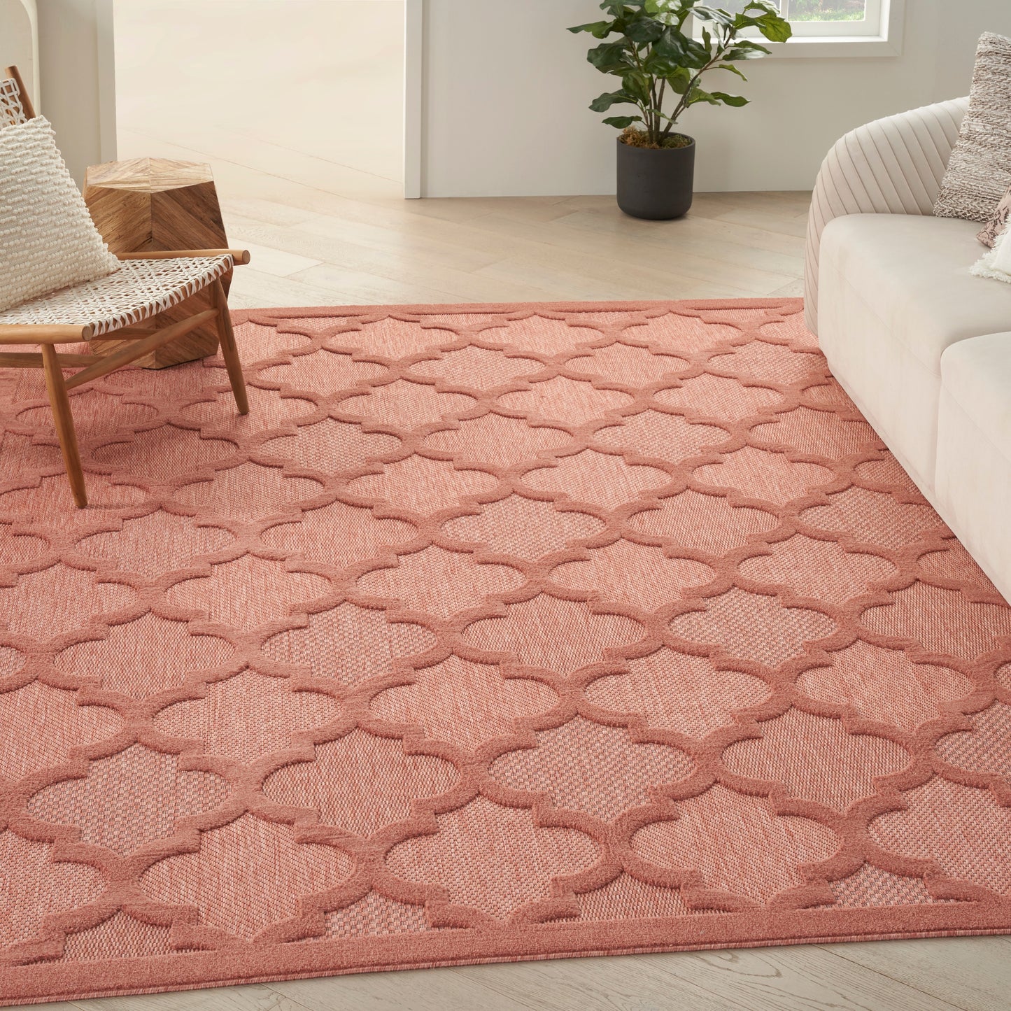 Coral woven area rug with repeating quatrefoil trellis pattern; living room scene with beige sofa and woven chair.