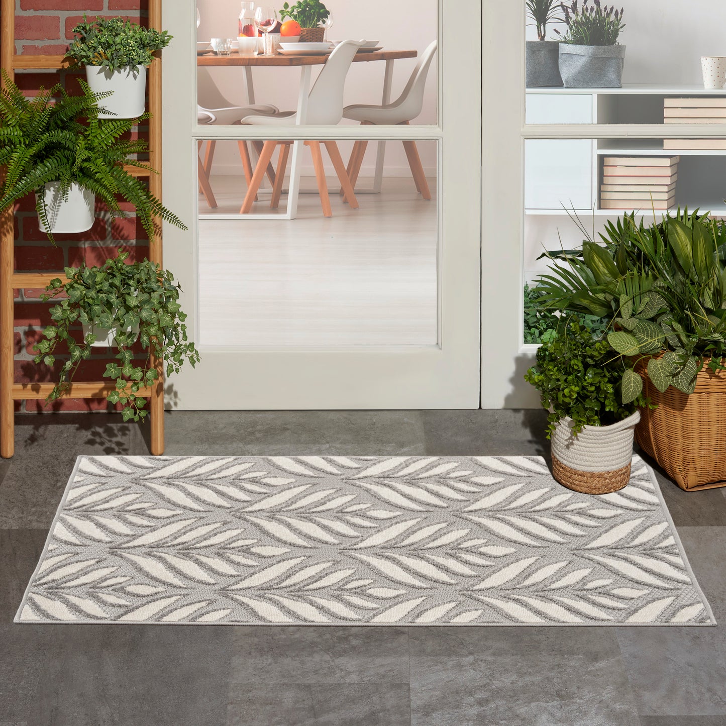 Rectangular grey and cream leaf-pattern outdoor rug at doorway, flanked by potted plants, leading to dining room.