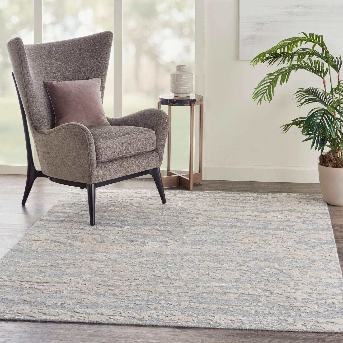 6' x 8' beige-gray rug in a living room, with a gray wingback chair with black legs, pink cushion, round side table, and a potted plant.