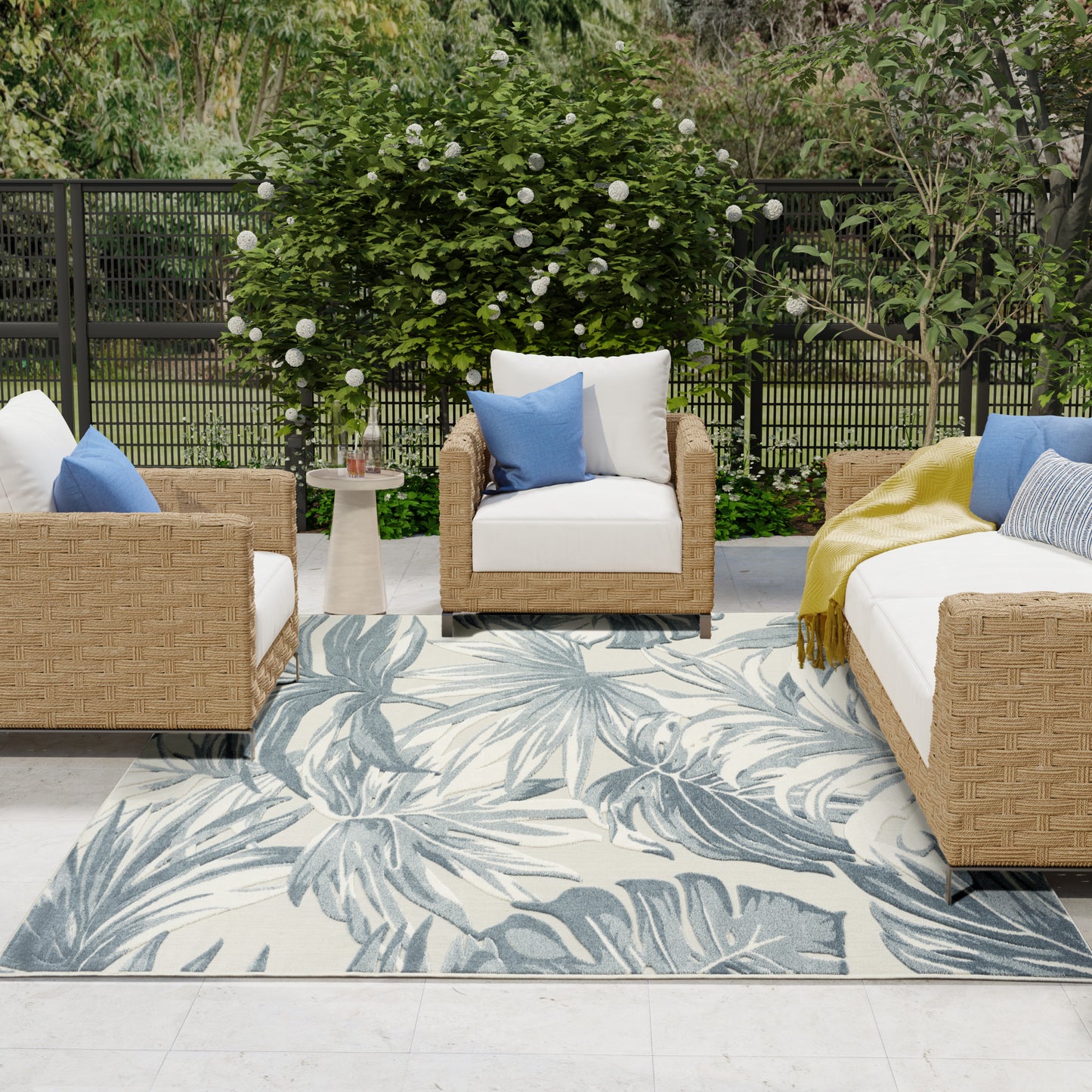 Outdoor wicker seating set with white cushions, blue accent pillows, palm-leaf rug, round side table, lush garden backdrop.