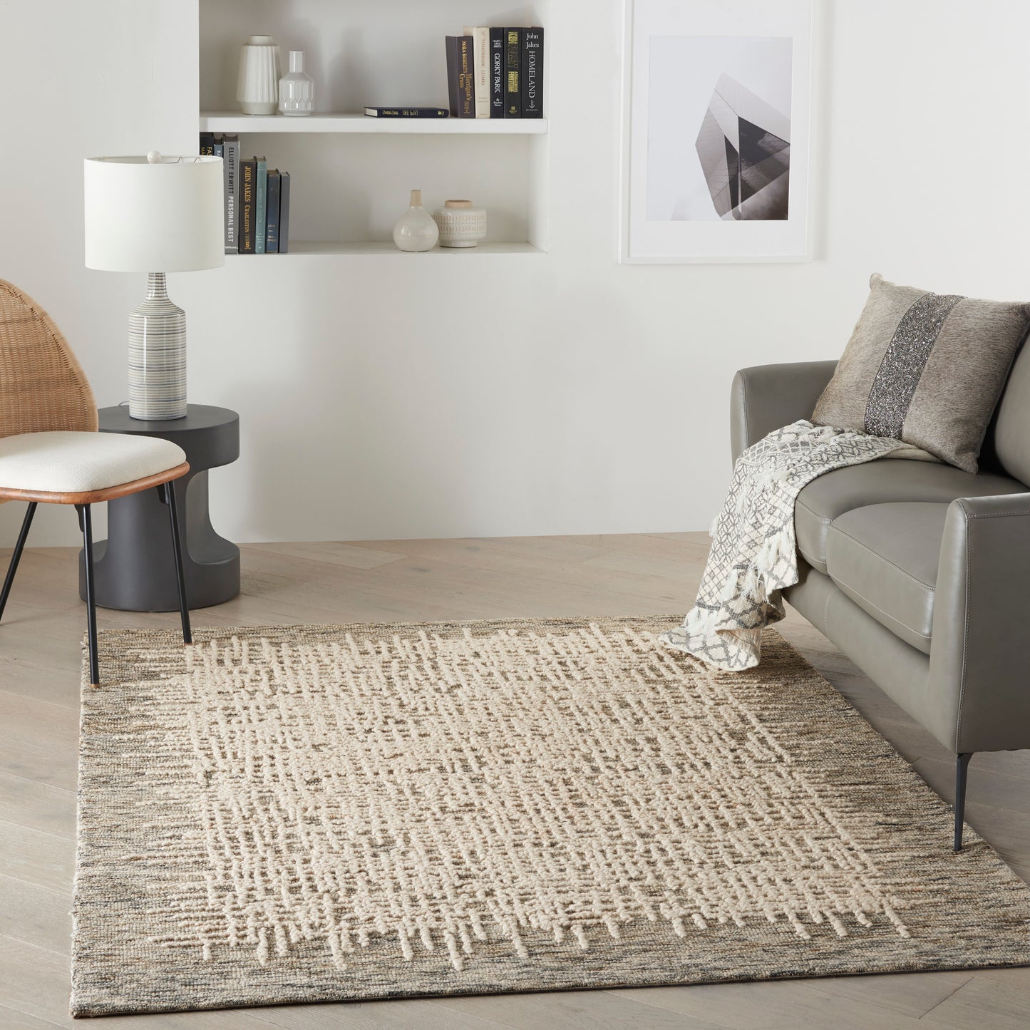 Neutral living room with beige woven rug, gray leather sofa, wicker chair, white shelves, and abstract wall art.