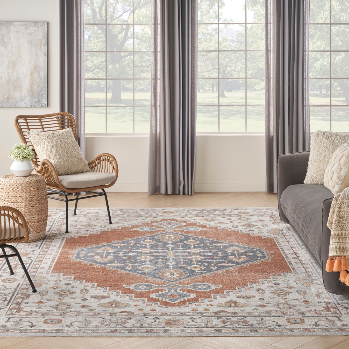 Living room with blue and orange patterned area rug, wicker chair and round side table, gray sofa, and large windows with gray drapes.
