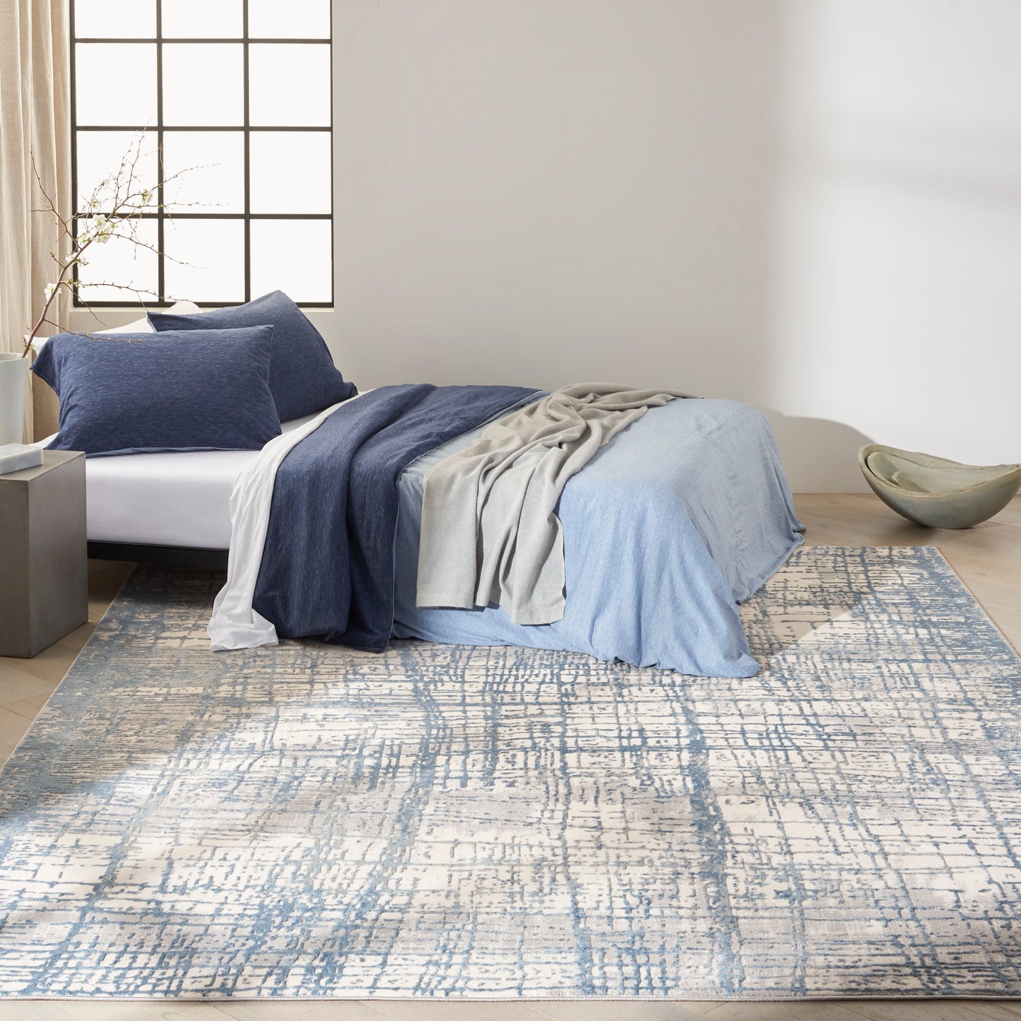 Low-profile bed with blue linens, gray throw and navy pillows on a light beige rug with blue abstract pattern.