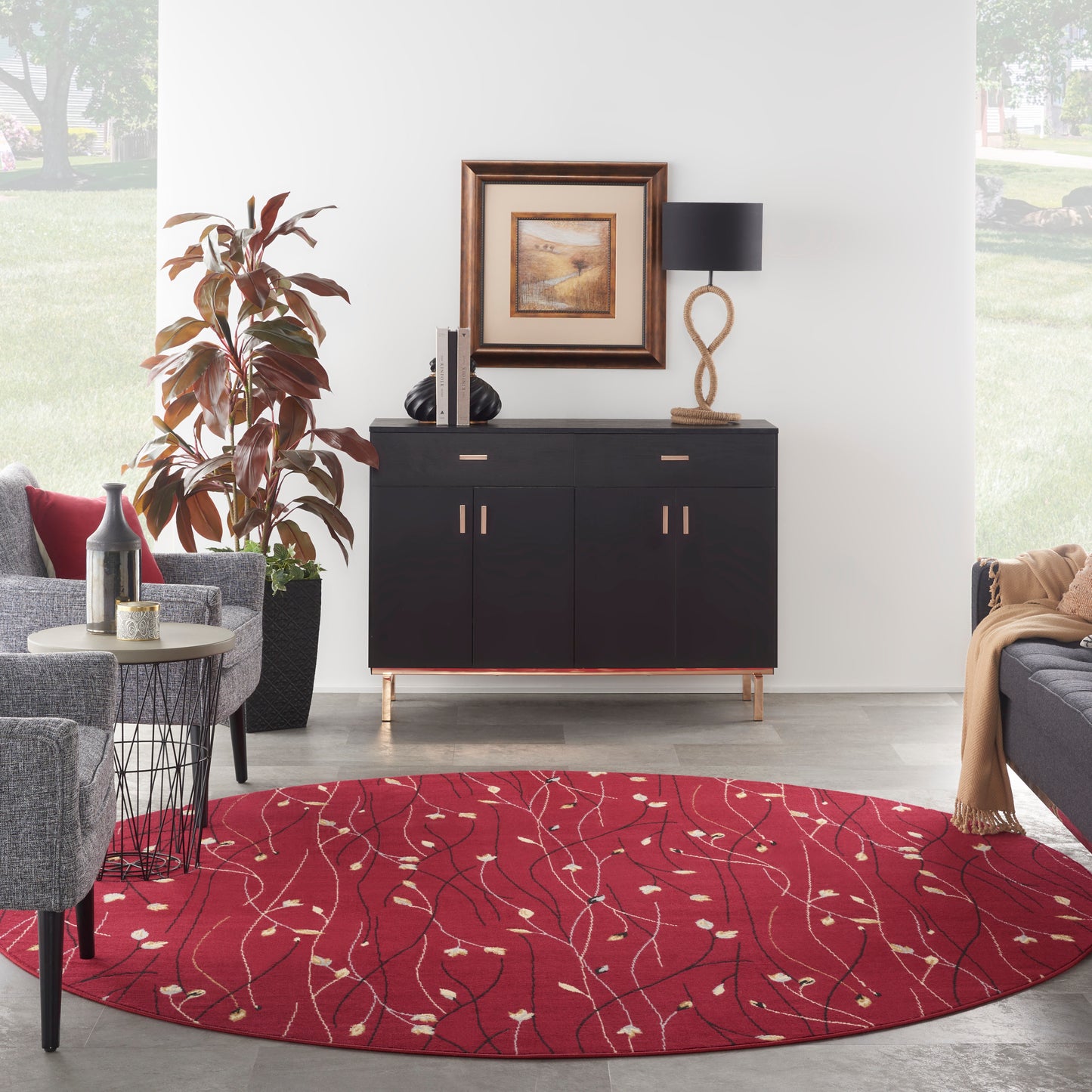 Round red rug with white leaf pattern under a black sideboard; framed art, rope lamp, gray seating and plant.