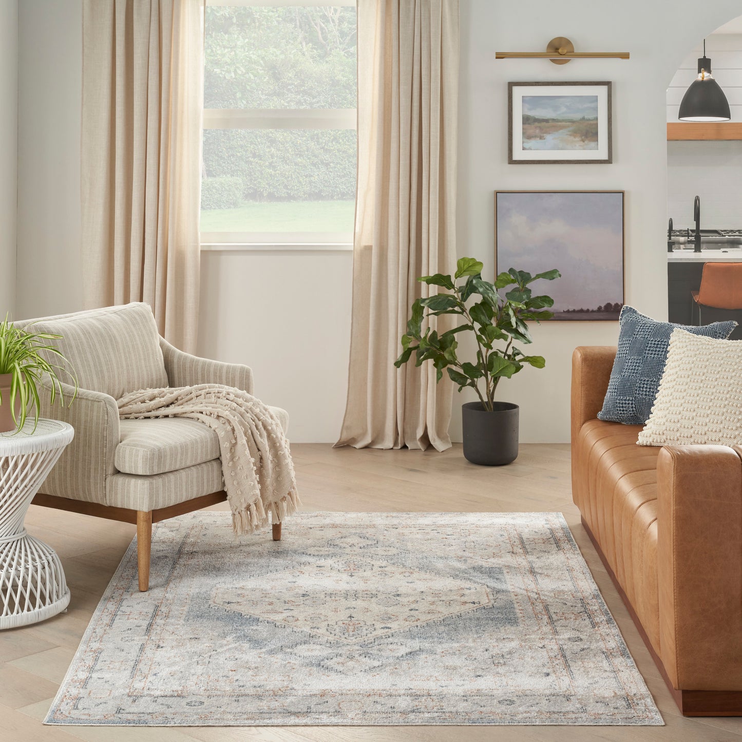 Cozy living room with light gray armchair, knit beige throw, muted blue patterned rug, tan leather sofa, and potted plant
