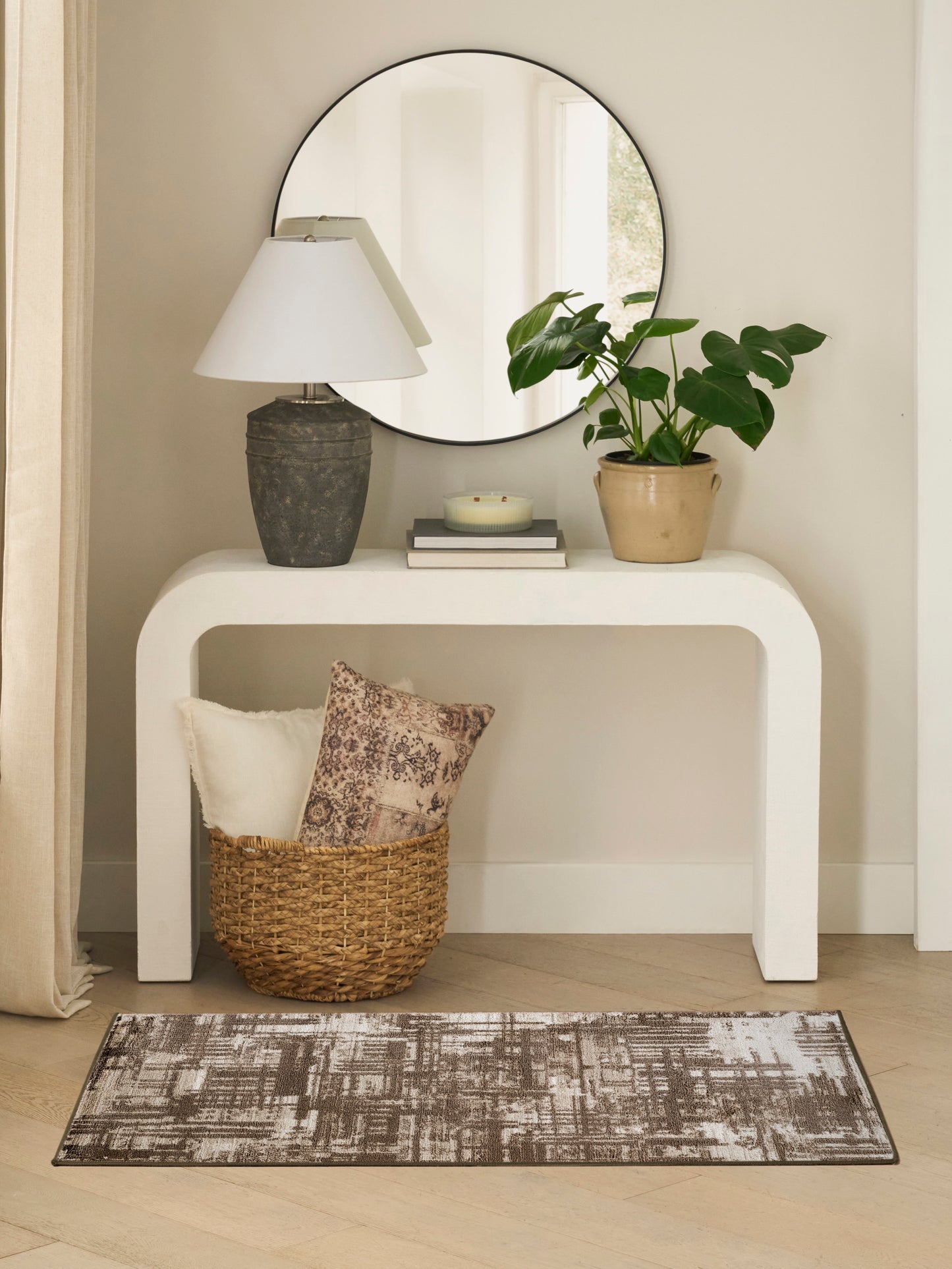 Neutral rug on the floor in a living-room vignette with a curved white console, round mirror, stone lamp, potted plant, candle on books, and a wicker basket with cushions.