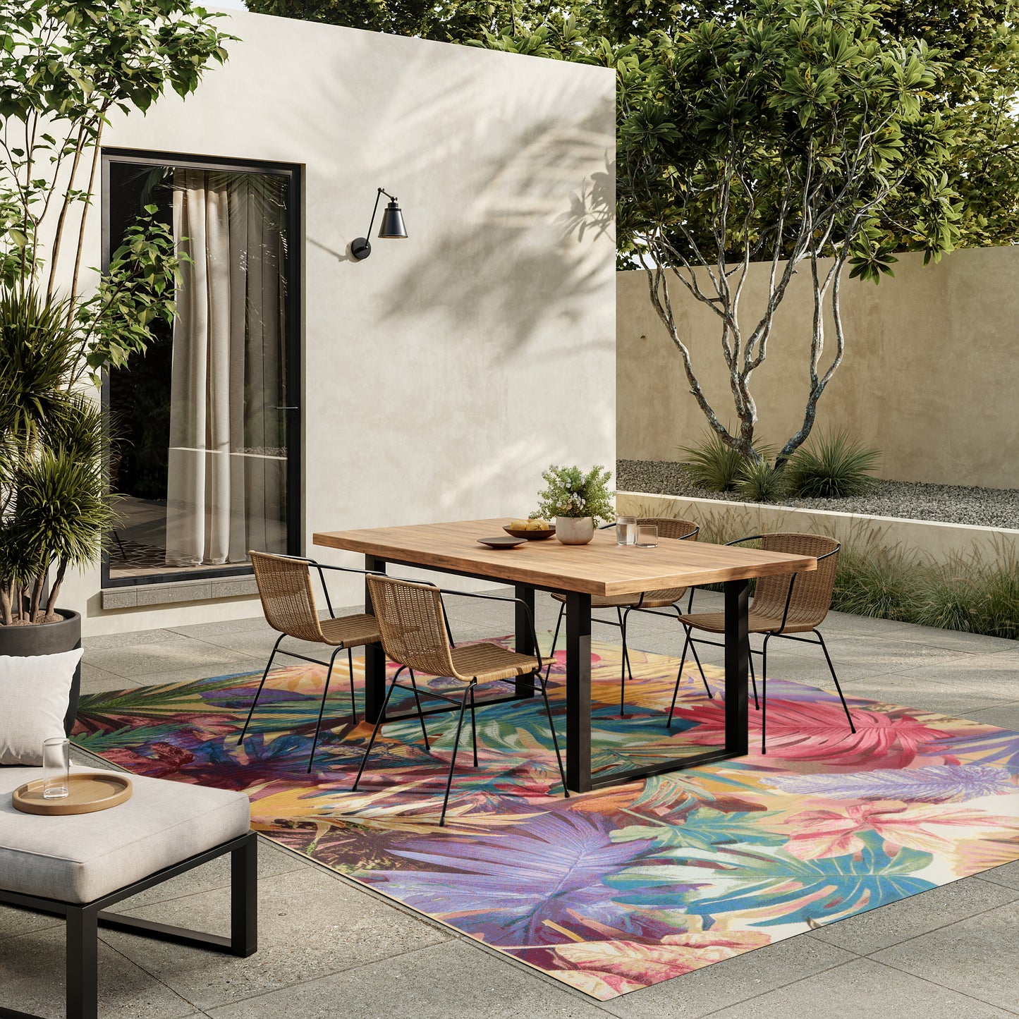 Outdoor dining area with a rectangular wooden table, six wicker chairs, a colorful tropical rug, beige stucco wall, and surrounding greenery.