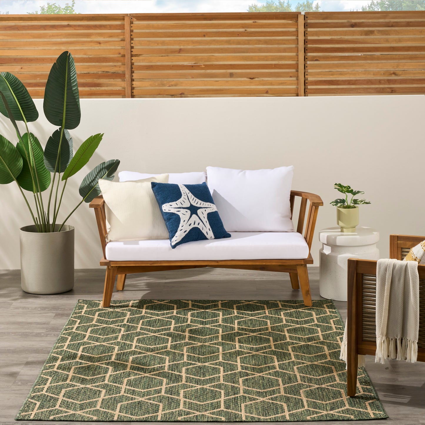 Wood outdoor sofa with white cushions and blue starfish pillow on a green geometric rug amid potted plants.