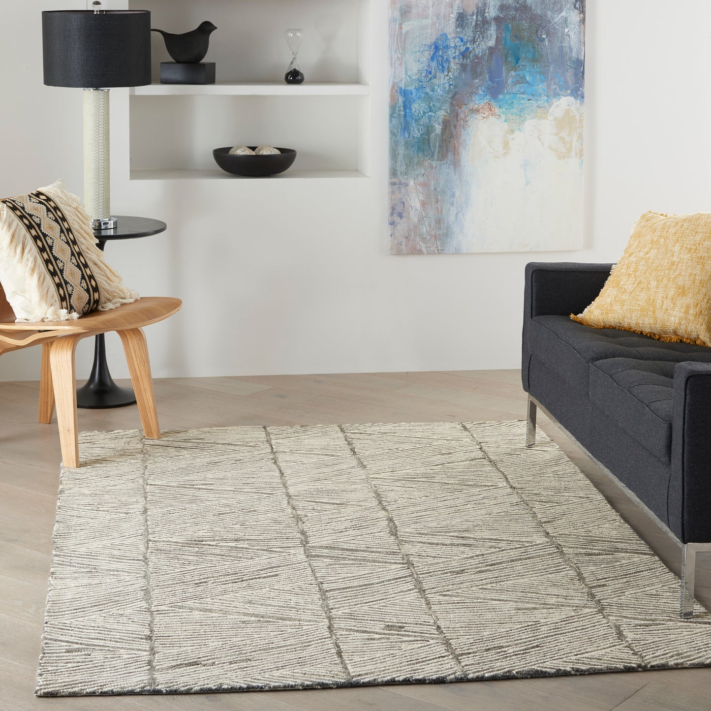 Modern living room with dark gray sofa, yellow throw pillow, beige rug, light wood chair, black lamp, and blue painting.