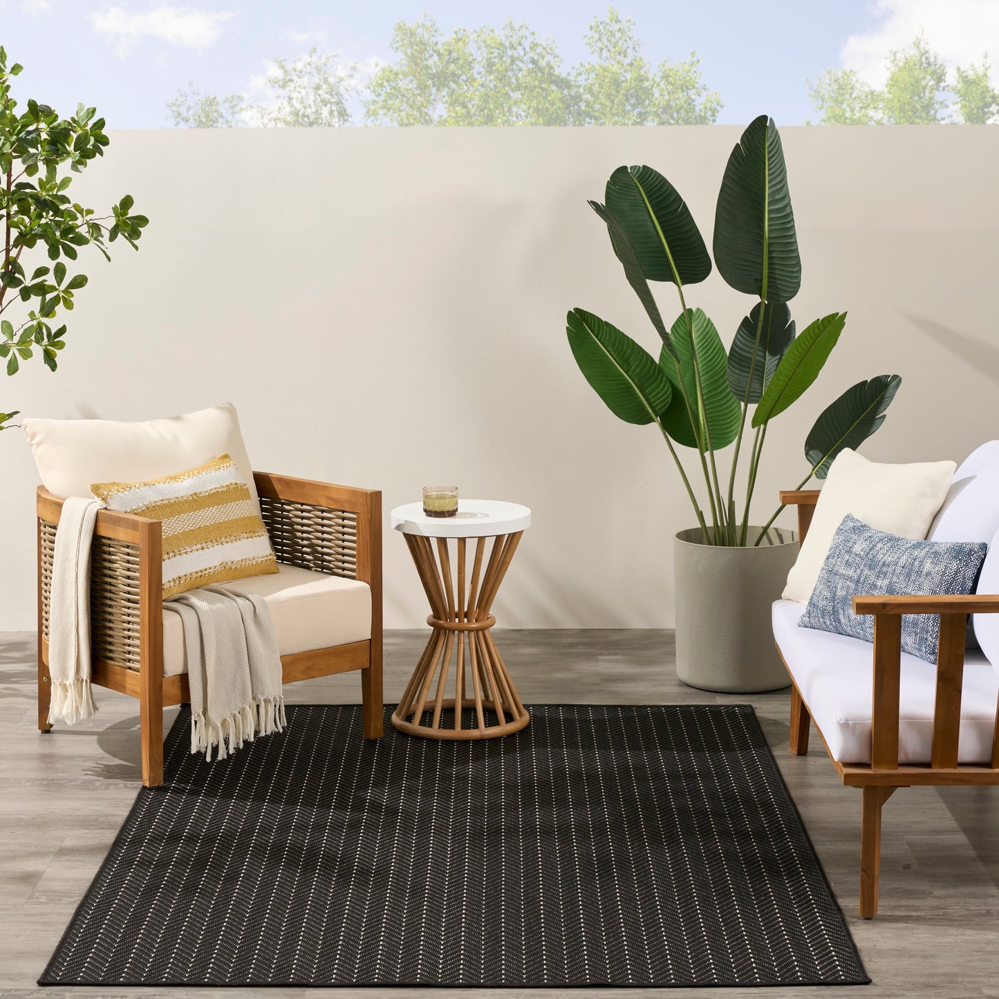 Outdoor patio scene with a black rug, wooden-frame armchairs with white cushions, a beige throw, a white side table, and a large tropical plant.
