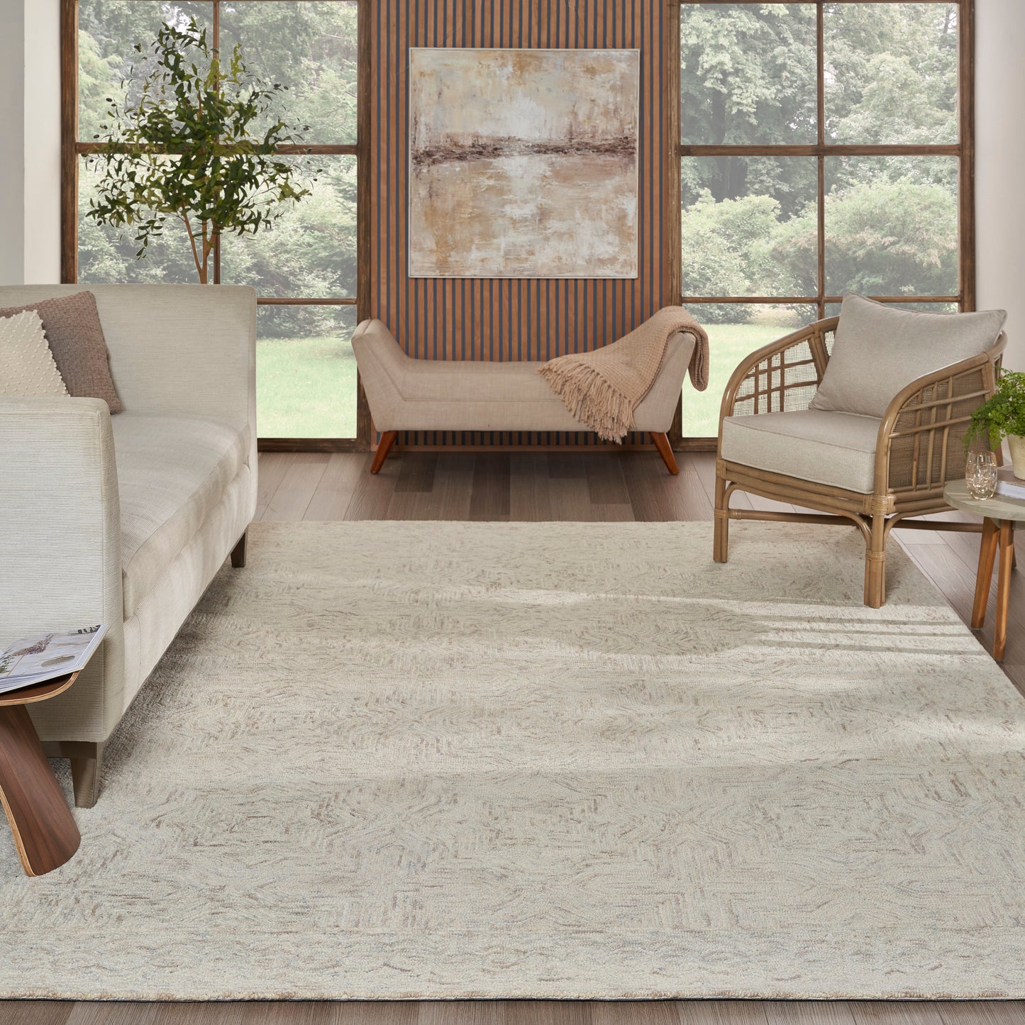Cream sofa and woven rattan chair in bright living room; neutral rug, wood panel wall, abstract art, large windows, greenery.