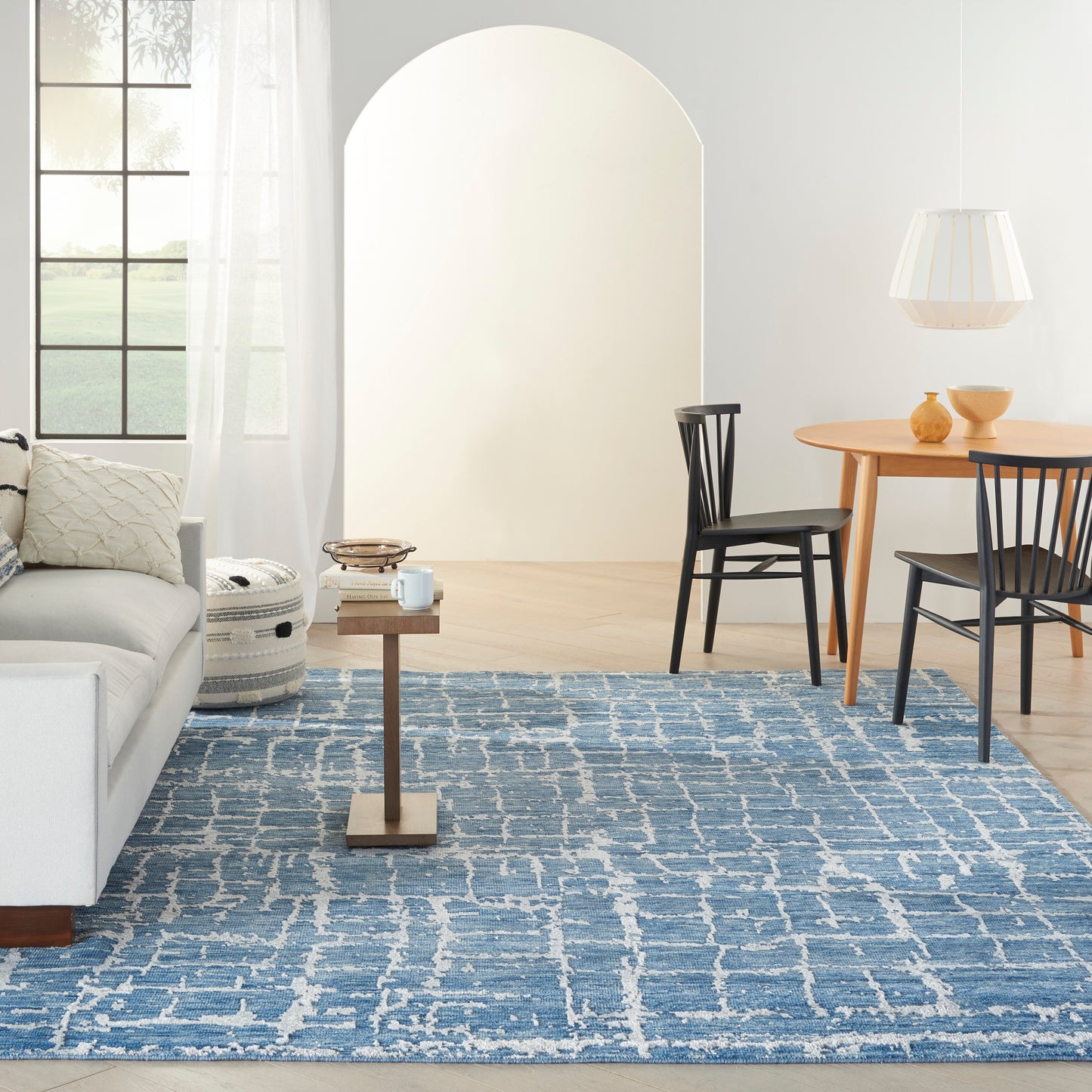 Bright airy living space with blue woven rug, light gray sofa, wooden side table, round dining table, black chairs, white pendant lamp, arched doorway.