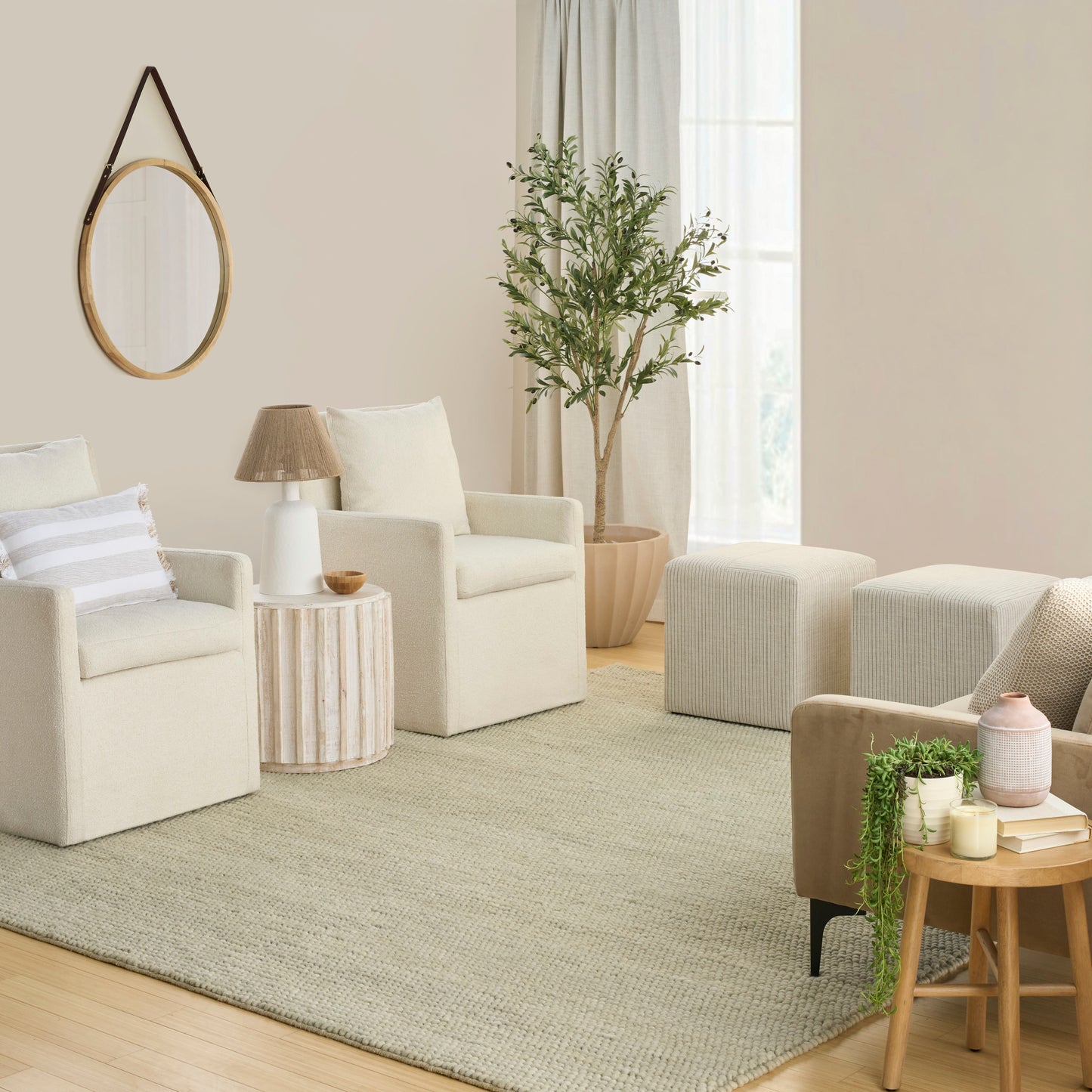Cozy neutral living room with cream armchairs, round wooden mirror, striped side table, potted plant, and beige rug.