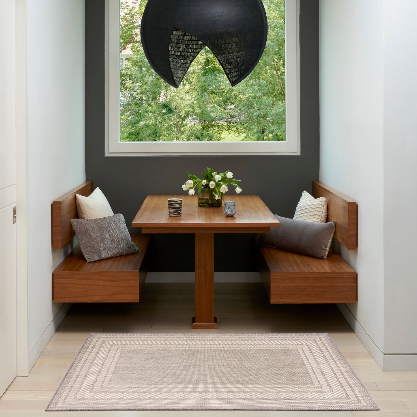 Cozy corner dining set with wood benches, rectangular table, black curved pendant, vase of white flowers, gray wall and rug