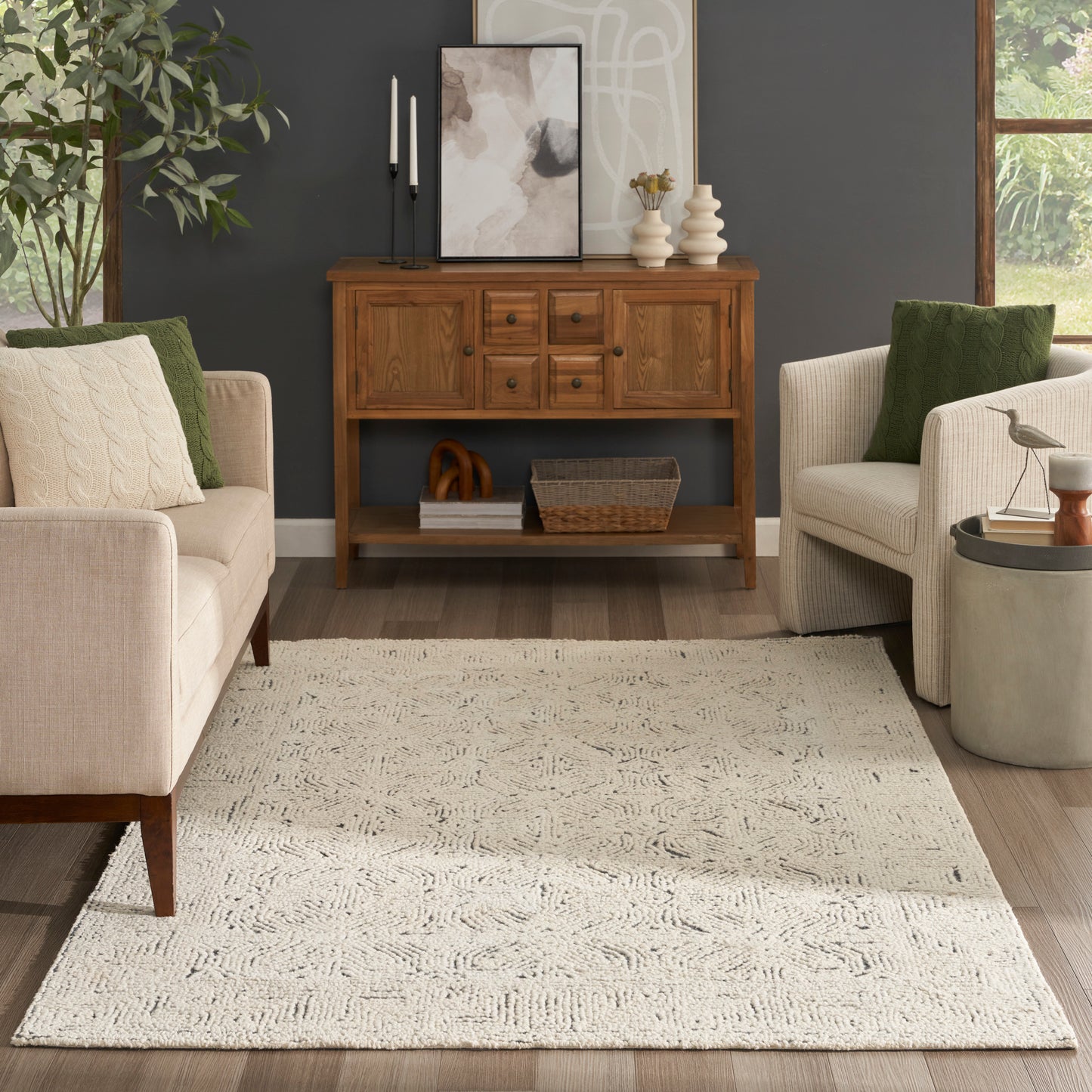 Wooden sideboard with drawers, beige fabric armchairs, cream textured rug, plant, dark gray wall, and abstract framed art.