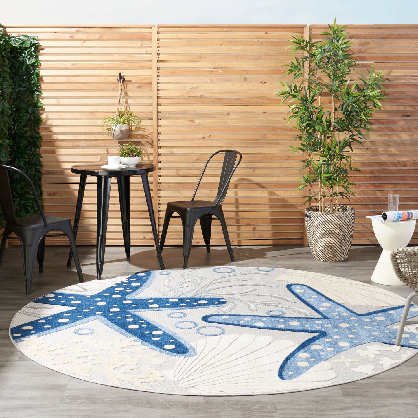Outdoor patio scene with a round blue starfish rug, black metal chairs, a small table, wooden slat fence and potted plants.