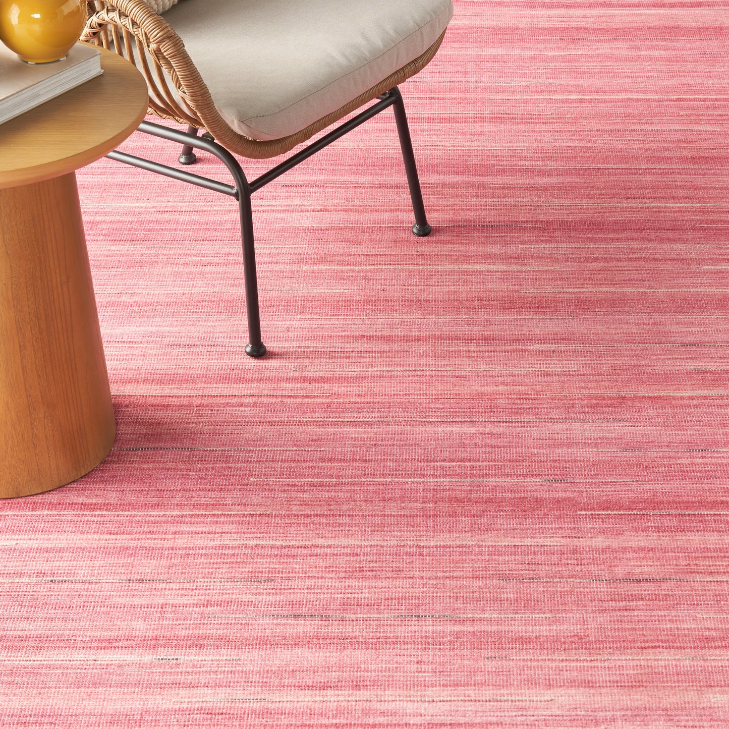 Pink striped flatweave rug, beige cushioned rattan chair & round wooden side table with an orange fruit.