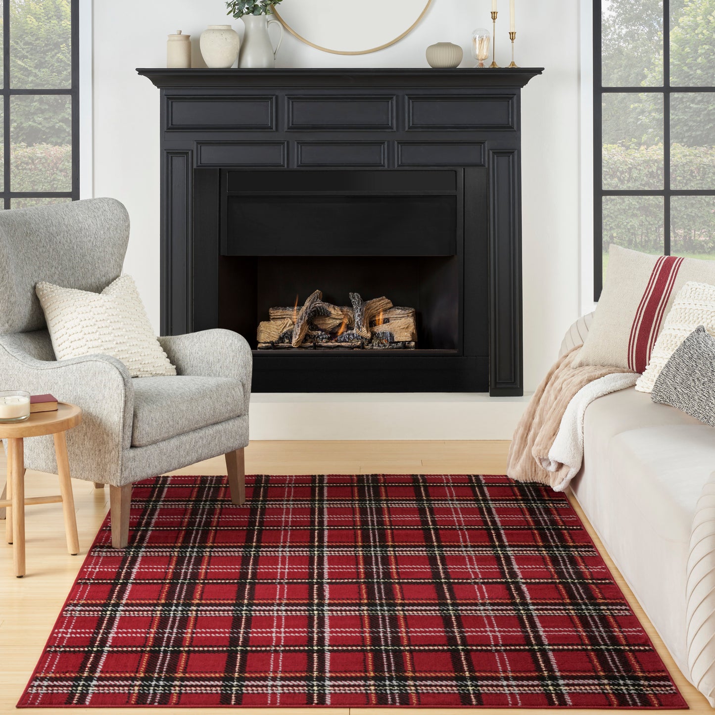 Black paneled fireplace with lit logs, gray armchair, red plaid rug, beige sofa and throw, round wood side table.