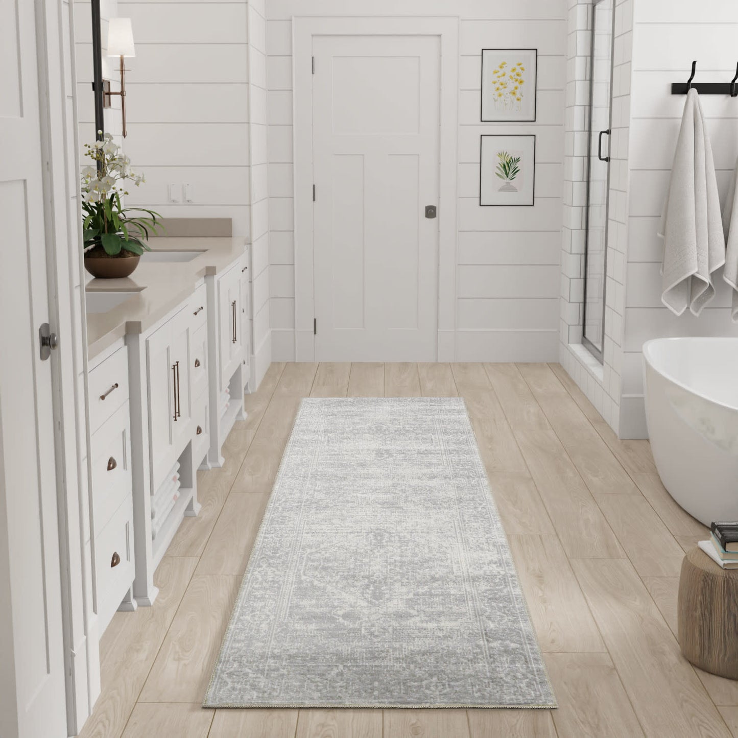 Bright white bathroom with light wood floor, gray runner, white vanity, botanical prints, glass shower, freestanding tub.