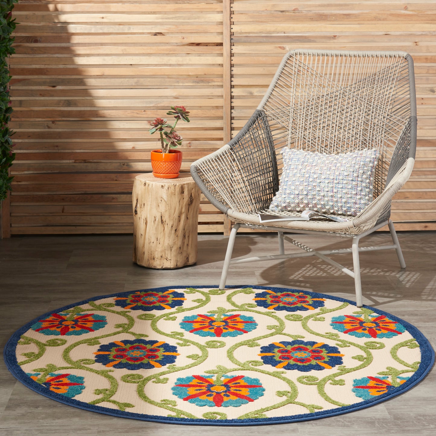 Round floral rug with blue border, beige base and bright orange, blue and green flowers, wicker lounge chair nearby.