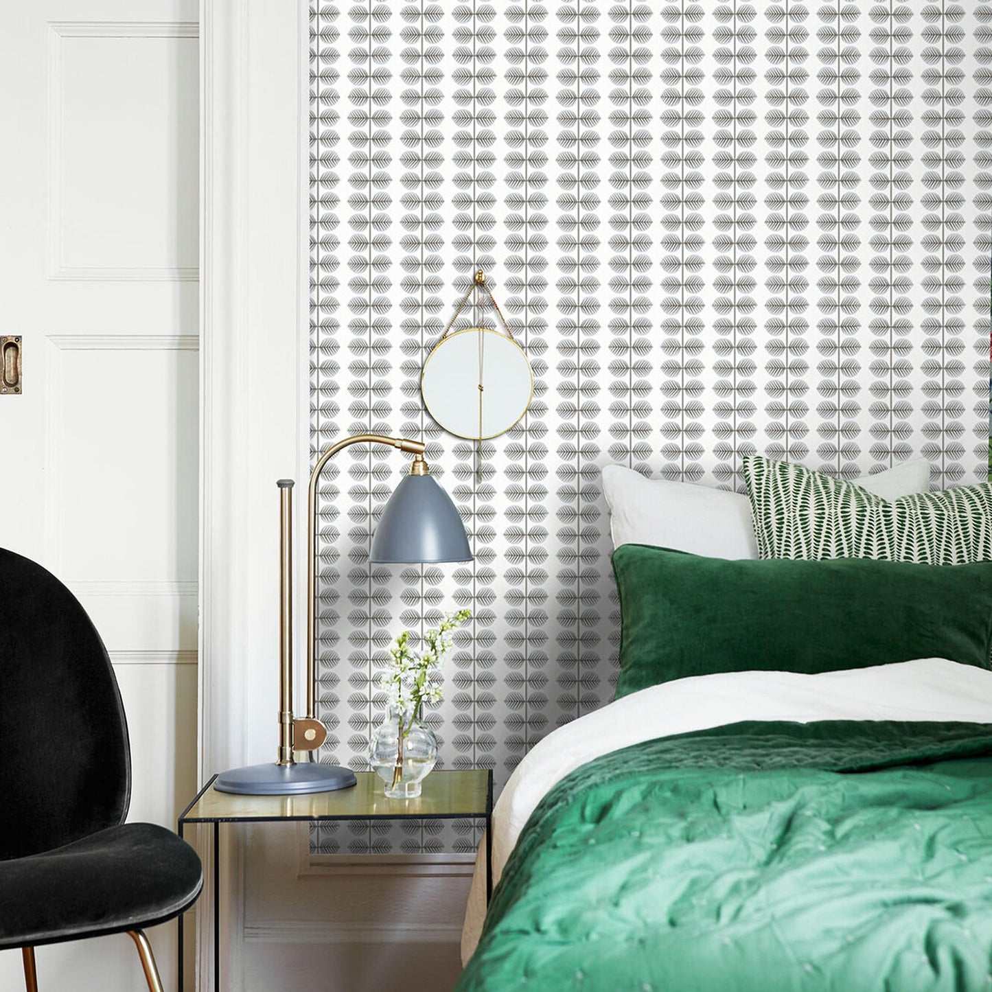 Modern bedroom featuring gray leaf-pattern wallpaper; visible decor includes a brass-blue lamp, a round gold clock, a glass vase, and a green duvet with white sheets.