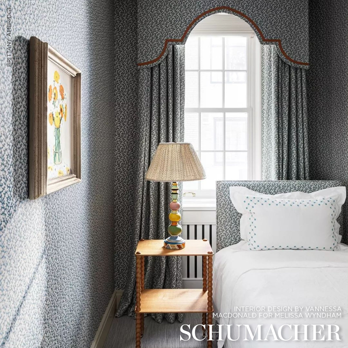 Blue and white wallpapered bedroom with arched window, blue drapes, wooden nightstand, stacked-sphere lamp, white bed.