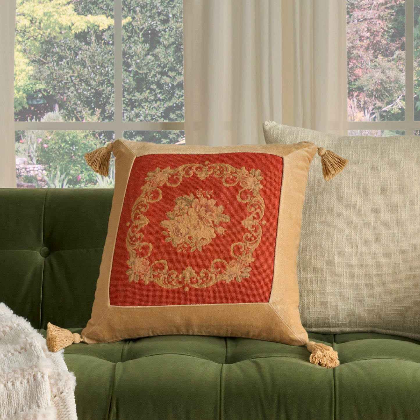 Decorative square throw pillow with red center and gold ornate design on beige border, tassel corners, on green tufted sofa.