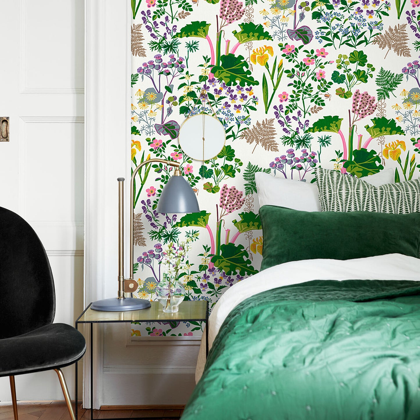 Bedroom with vivid botanical wallpaper, emerald bedding, brass bedside lamp, round mirror, & a black chair.