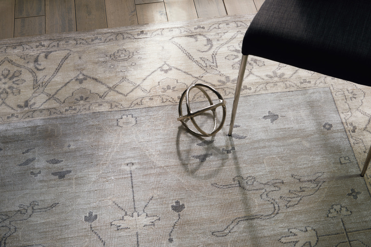 Light gray patterned area rug with floral motifs; a metallic geometric orb sculpture sits near a black chair with metal legs.