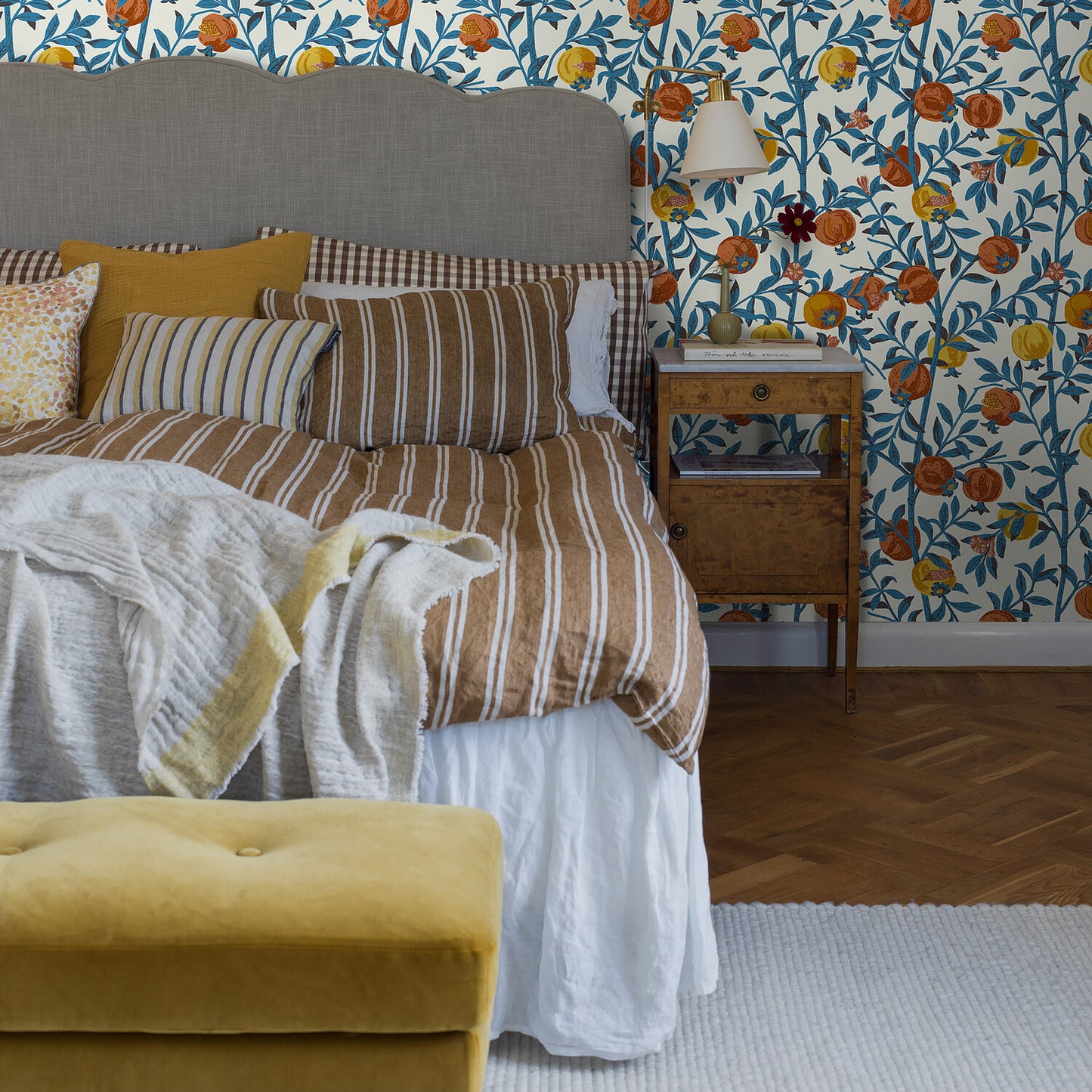 Blue floral wallpaper, gray headboard, brown striped bedding, mustard cushions, cream throw, yellow velvet ottoman.
