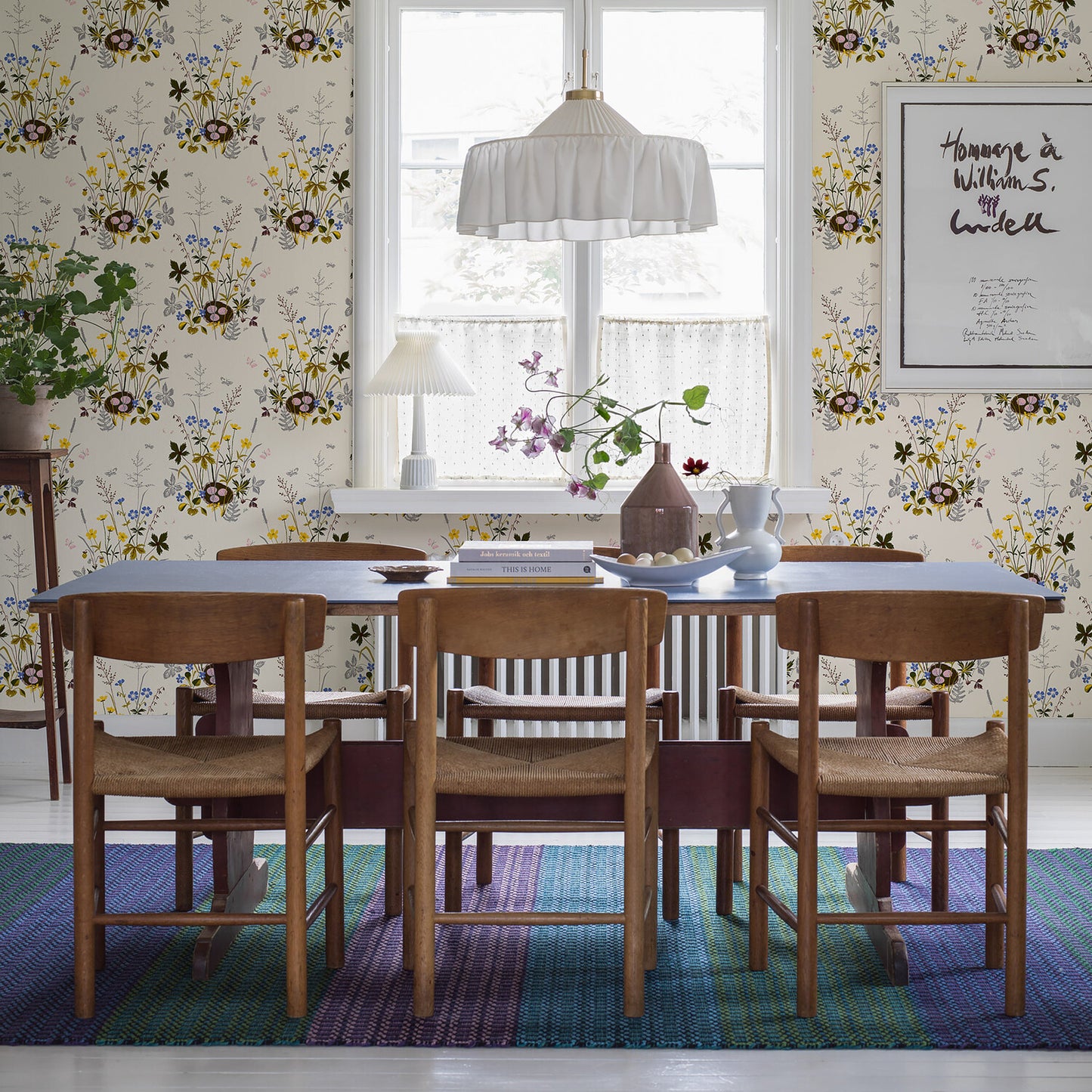 Mid-century wooden dining table with six chairs, floral wallpaper, blue rug, white pleated pendant lamp, plants.