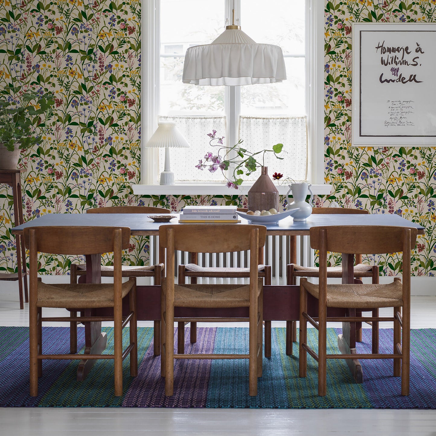 Mid-century wooden dining table with chairs, bold botanical wallpaper, blue striped rug, and white pendant light.
