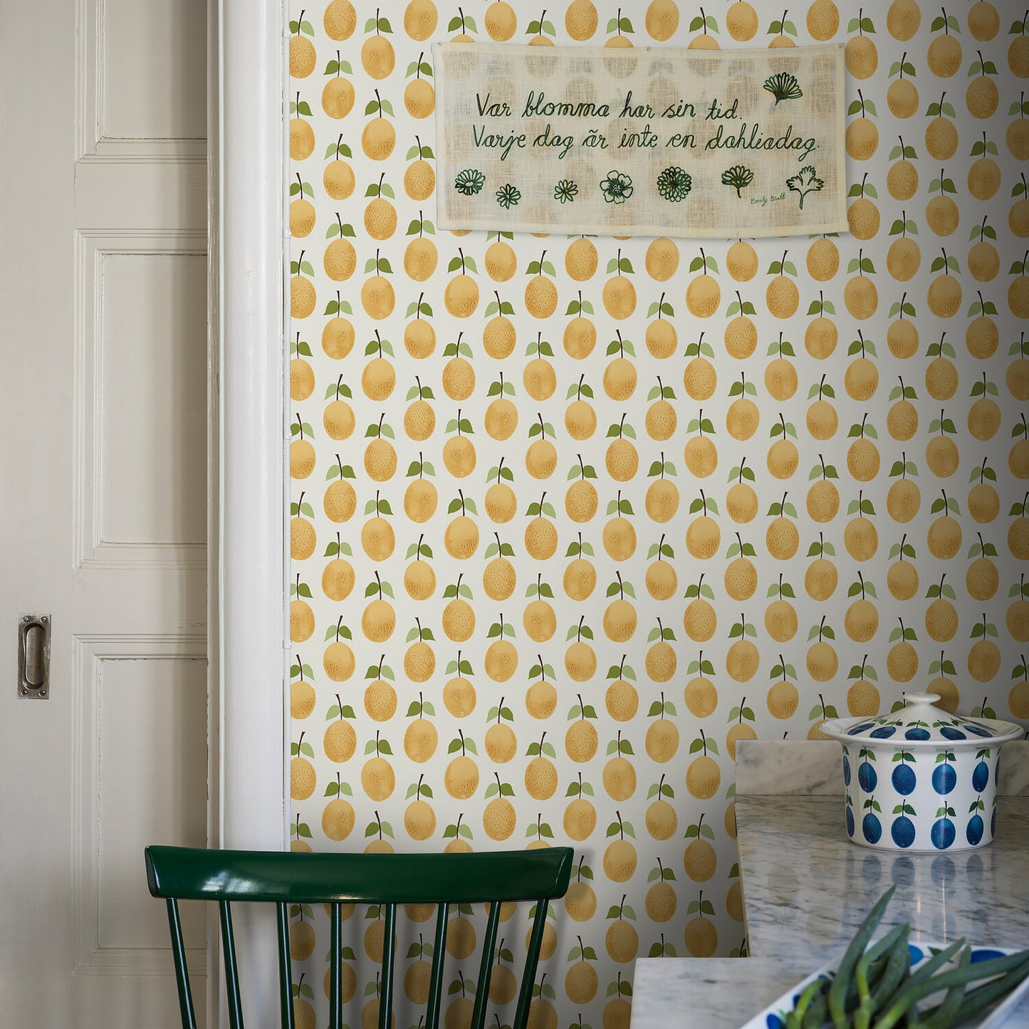Retro kitchen nook with lemon-pattern wallpaper, white door left, green chair, marble counter, and a blue-dotted ceramic jar.