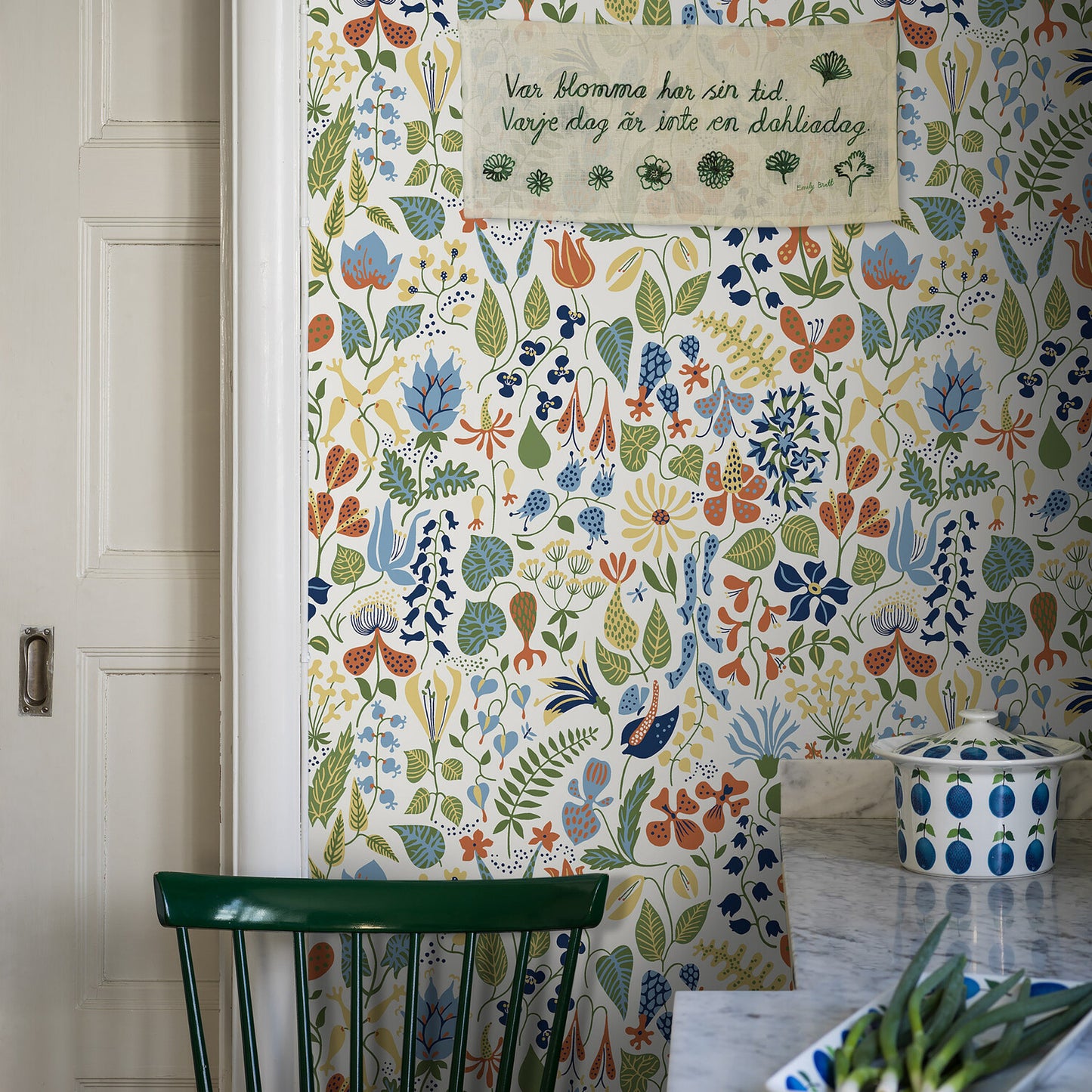 Colorful botanical wallpaper with leaves and flowers; white door left, green chair, marble counter, blue patterned jar, sign.