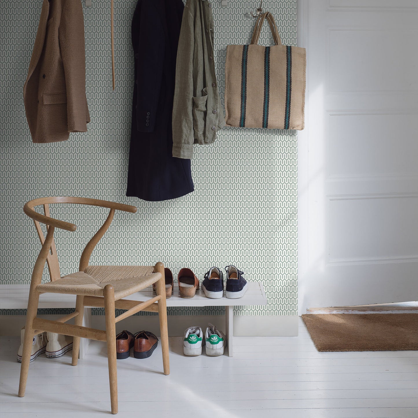Entryway with wallpaper, wooden chair, coats on hooks, beige striped tote, white bench and sneakers, light wood floor.