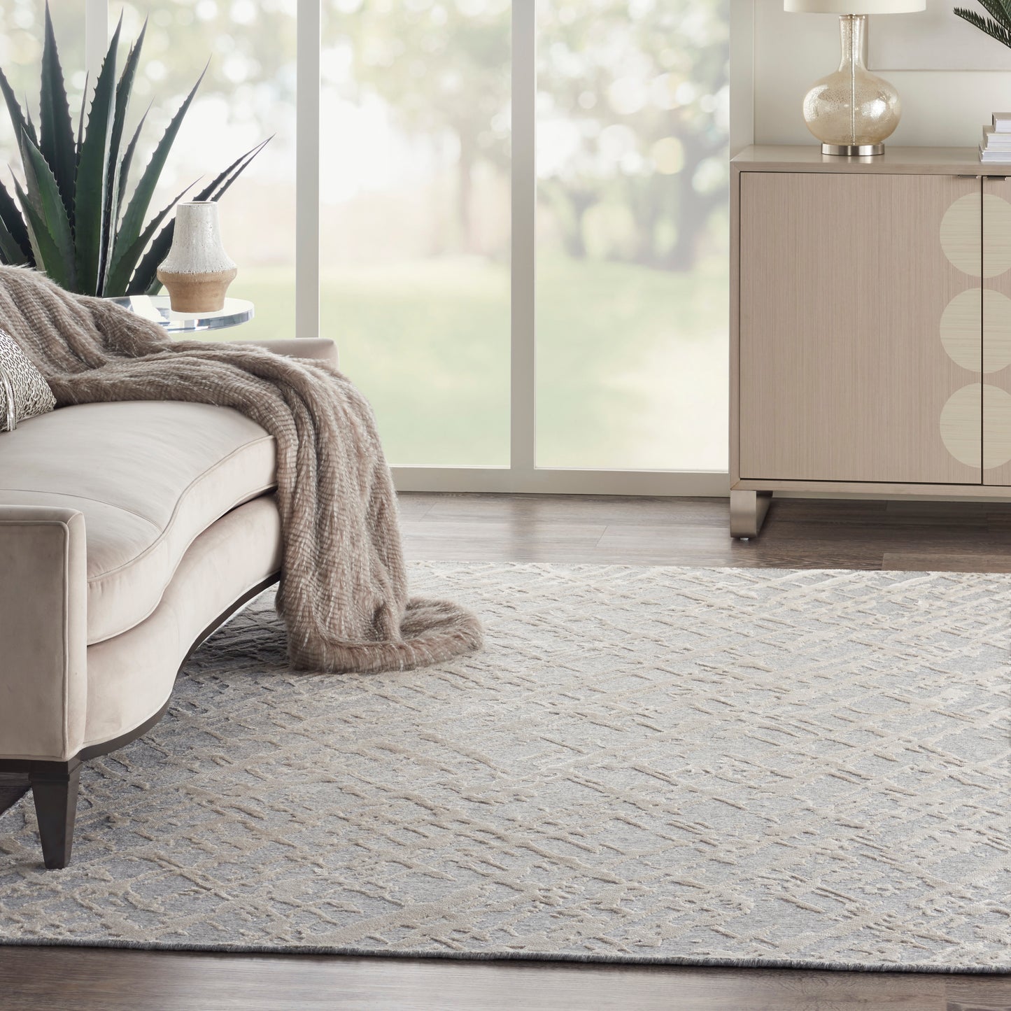 Beige sofa with taupe knit throw, light gray patterned rug, large plant, and light wood cabinet by sunny windows.