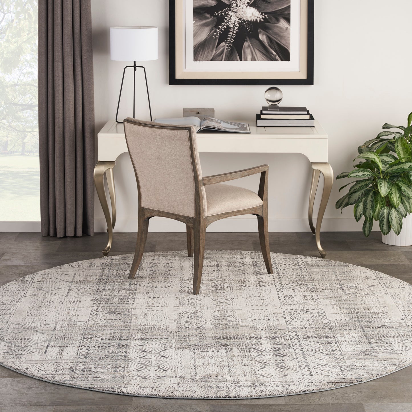 Beige upholstered desk chair with wooden frame at a white desk, round gray patterned rug, lamp, plant and framed art.