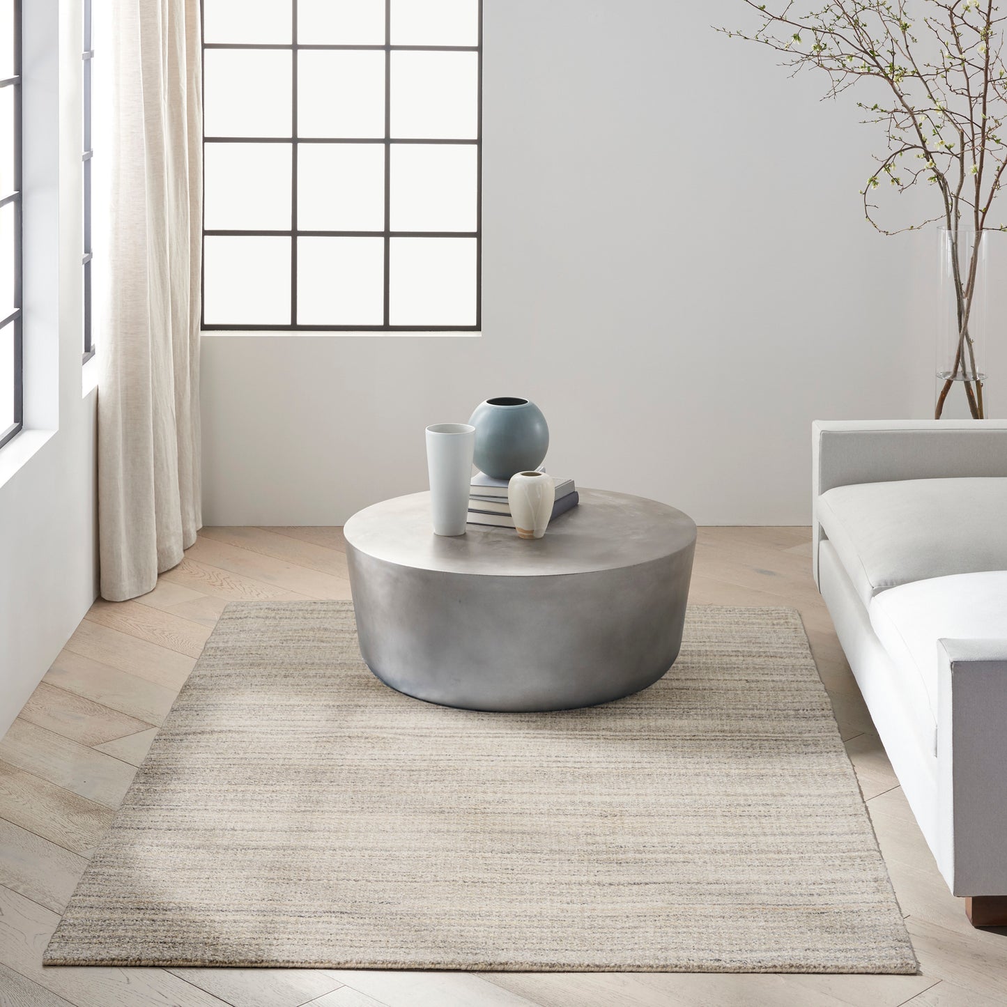 Minimalist living room with round concrete coffee table, ceramic vases, beige rug, grid-window, white sofa.