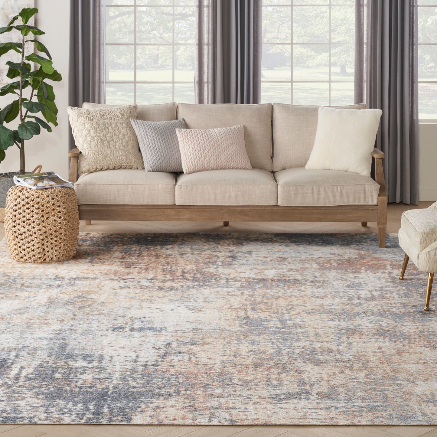 Beige fabric sofa with wooden frame, neutral knit pillows, woven rattan side table, blue-gray abstract rug, potted plant.