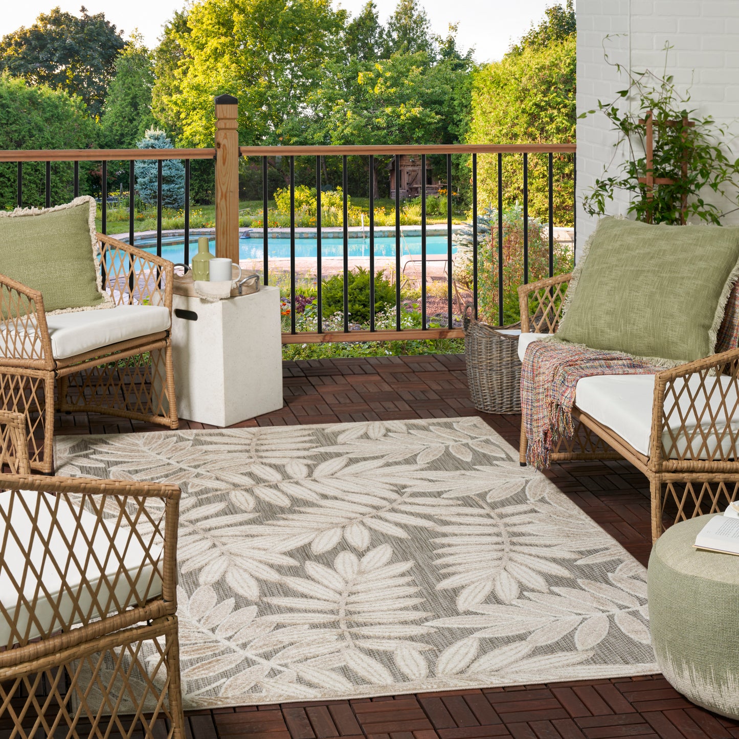 Leaf-pattern outdoor rug on a wooden deck, with outdoor wicker seating featuring beige cushions & green pillows; pool & greenery beyond.