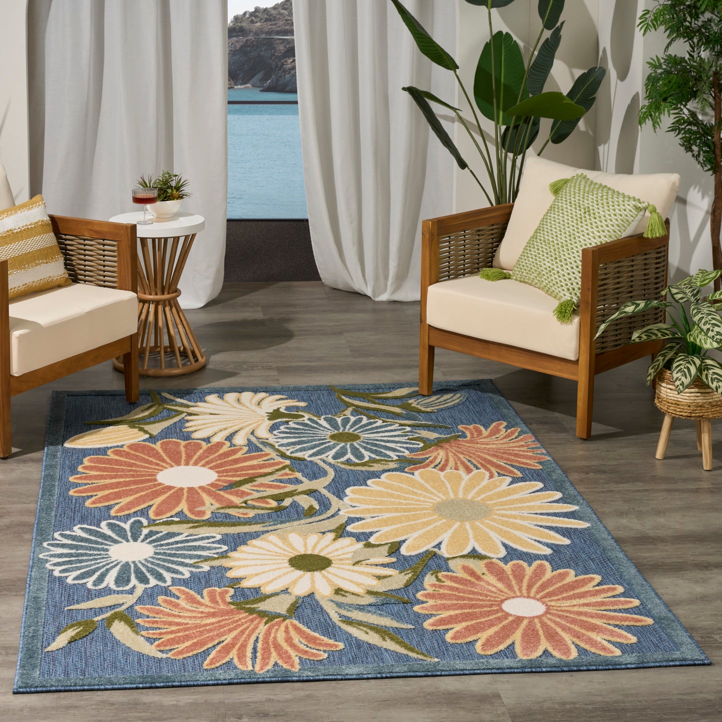 Blue floral rug with large daisies; wooden-framed wicker chairs with cream cushions; tropical plants & seaside view.