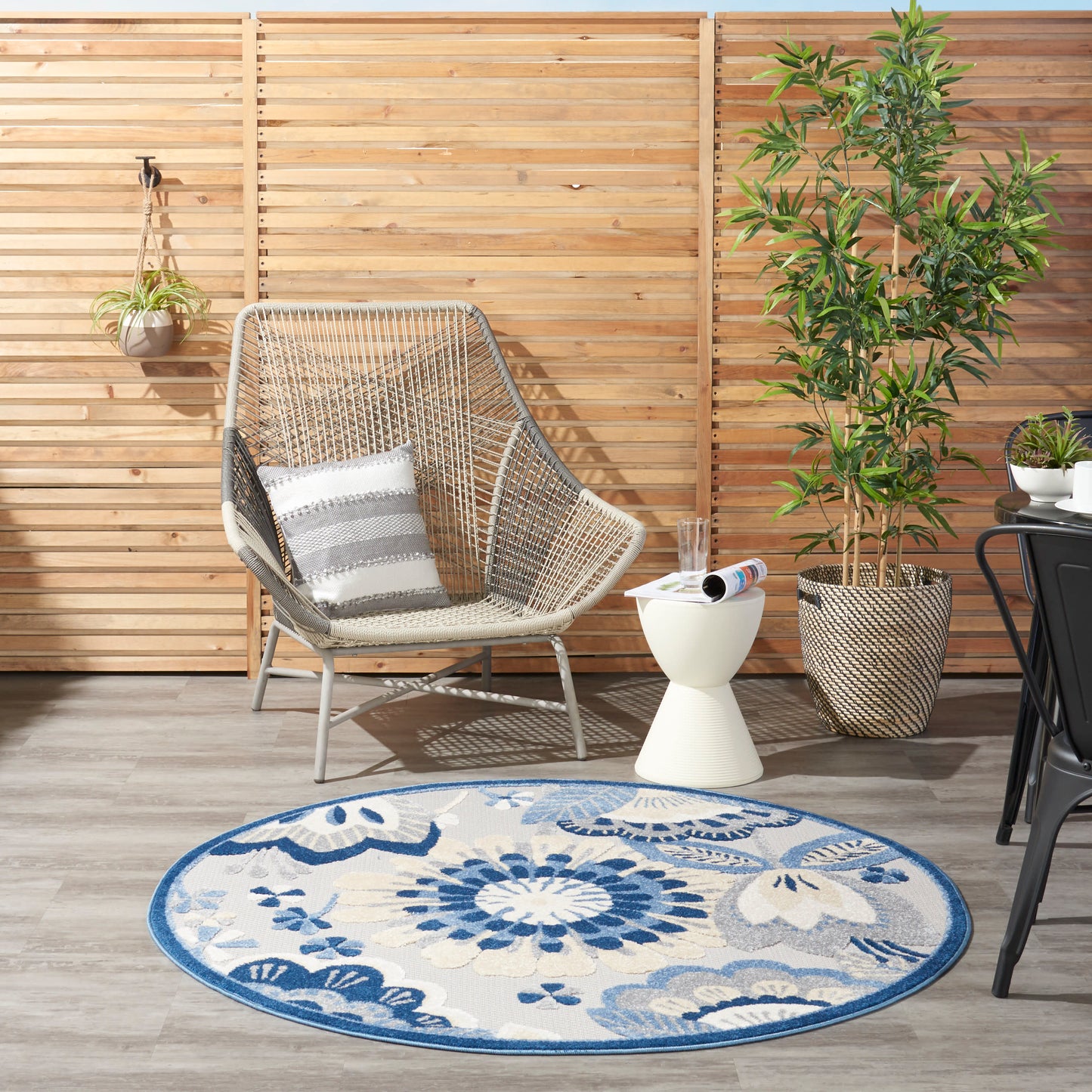 Outdoor patio with round blue floral rug, beige wicker chair with striped cushion, wooden slat fence, potted plants.