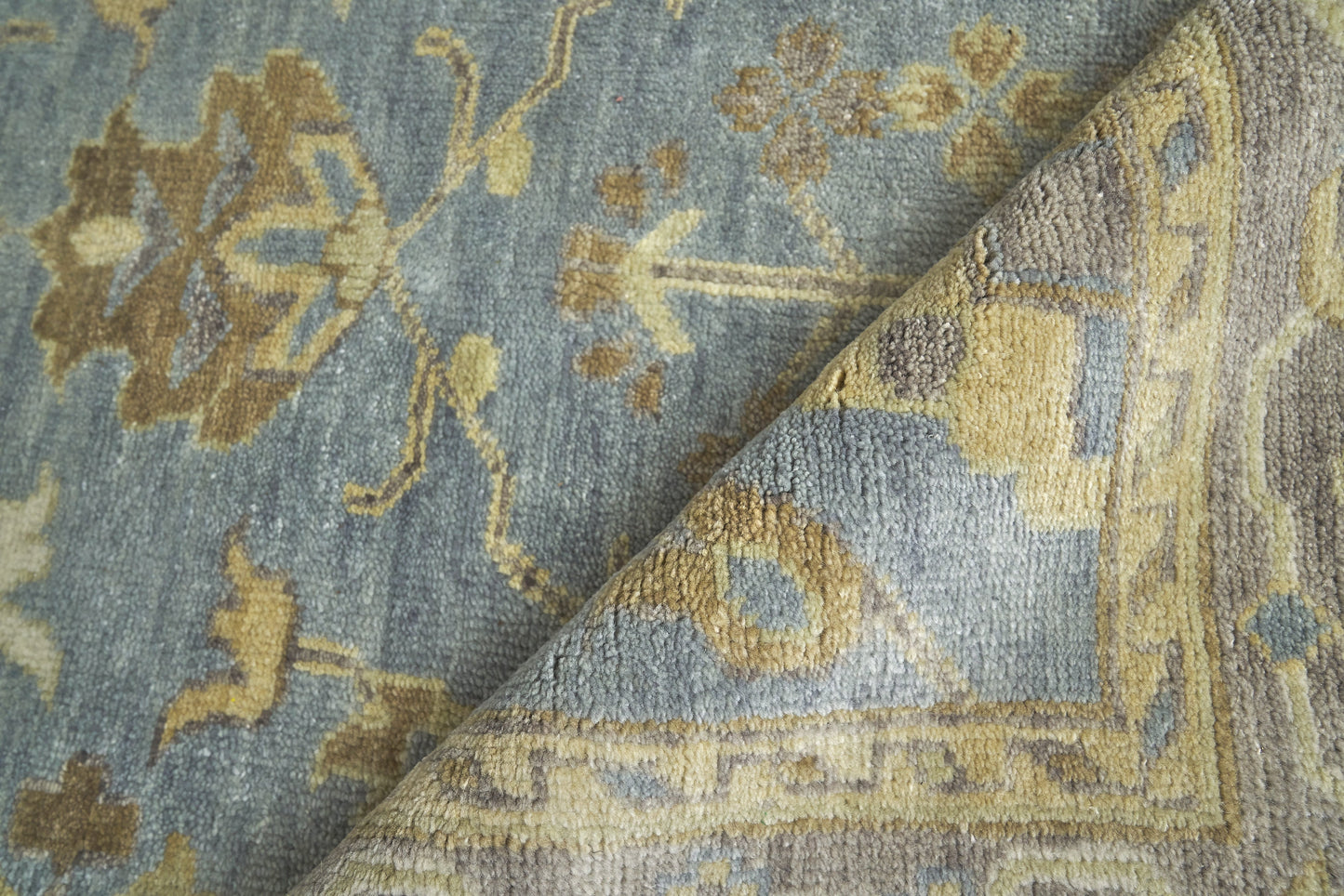 Blue-gray oriental area rug with mustard floral accents and geometric border; plush wool pile, folded corner.