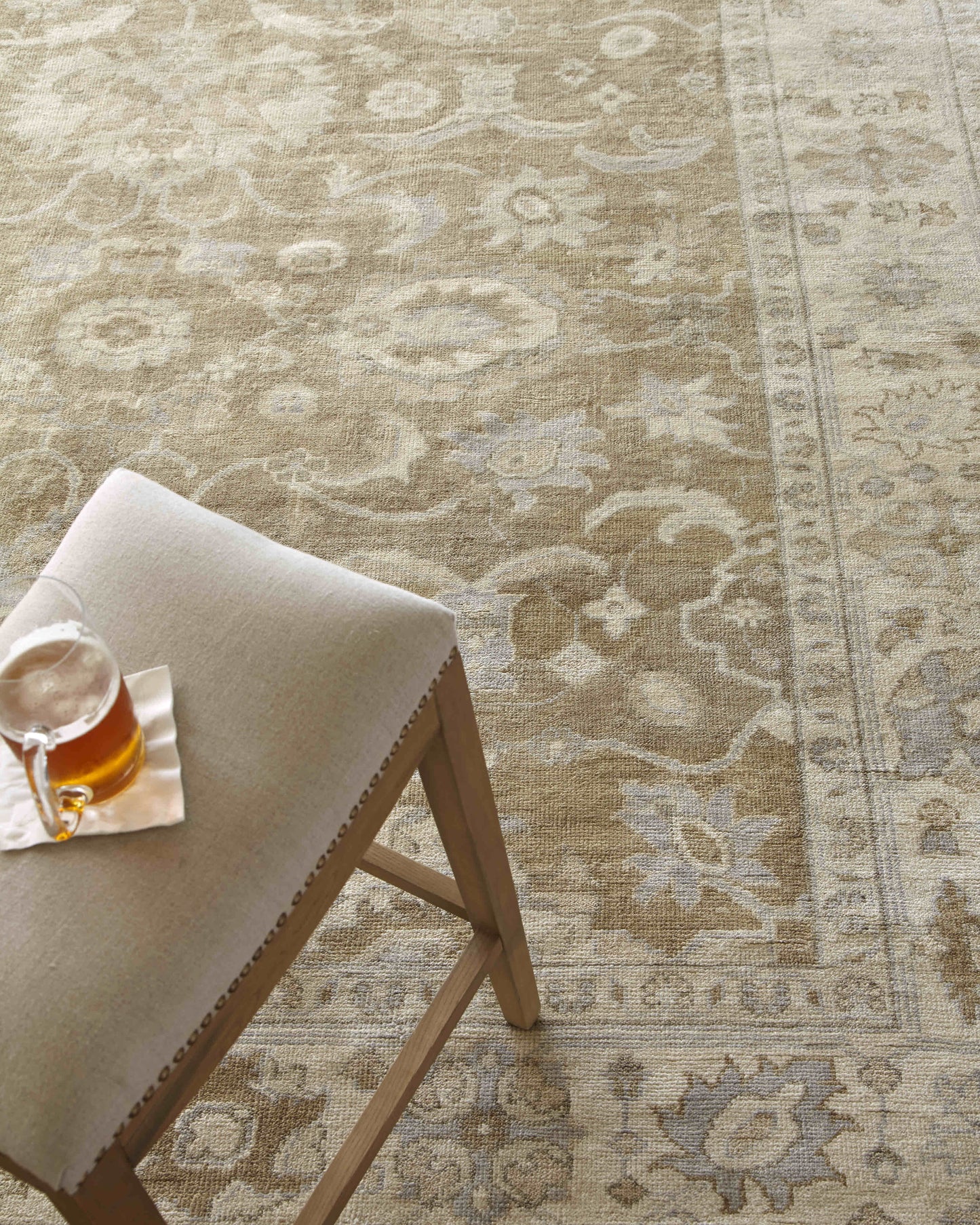 Beige floral-pattern area rug; nearby a light wood stool with a beige cushion and a glass mug of tea on a napkin.
