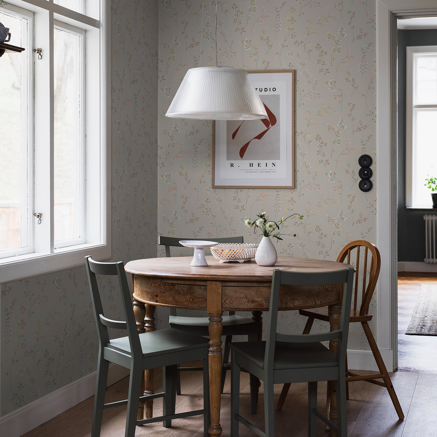 Round wooden dining table with turned legs, mixed gray-green and natural chairs, white pendant lamp, stone-pattern wallpaper on the wall.