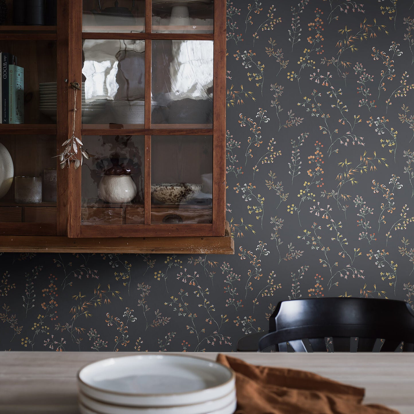 Cozy vintage kitchen corner: dark floral wallpaper, wooden glass-front cabinet with dishes, and a table setting with a white plate and brown napkin.