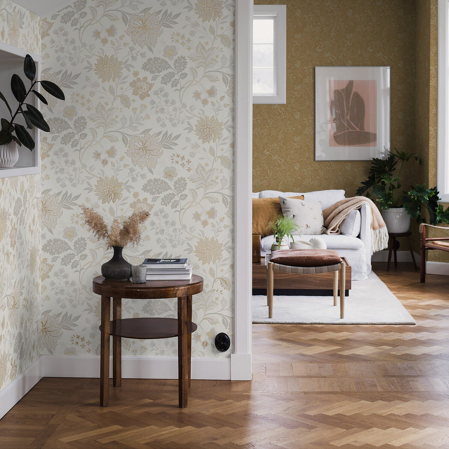 Cozy living room: beige floral wallpaper, round wood side table with dried flowers, herringbone floor, and a sofa with throw.