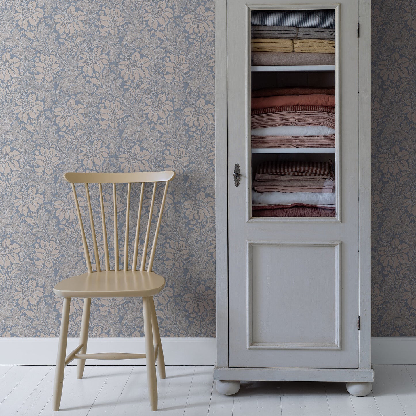 Creamy yellow wooden spindle-back chair beside a white glass-front cabinet with folded blankets; vintage floral wallpaper.